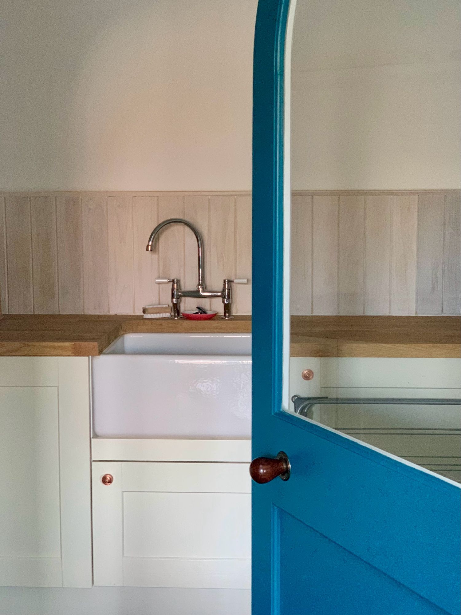 Picture of a bunker, Belfast sink, curved tap and door.  The bunker is wooden, the sink is white, the cupboard doors are cream and the room door is teal blue.  The photo is taken through the open door, the light is diffuse and soft giving a painted feel.  There is a tiny orange dish under the tap which brings a tiny bit vital pop of colour contrast to the cream and dominating teal.