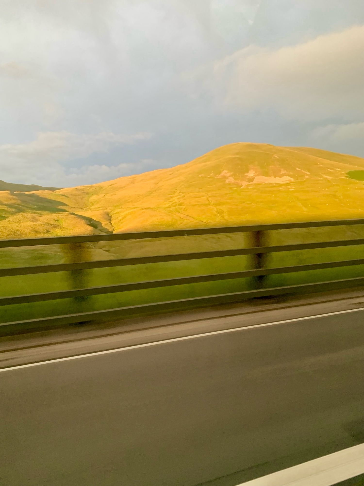 Picture taken on the move, you can see road, blurry fencing, a hill being hit by the sun - its lime green.  The sky is a domineering pale grey as rain is threatening.