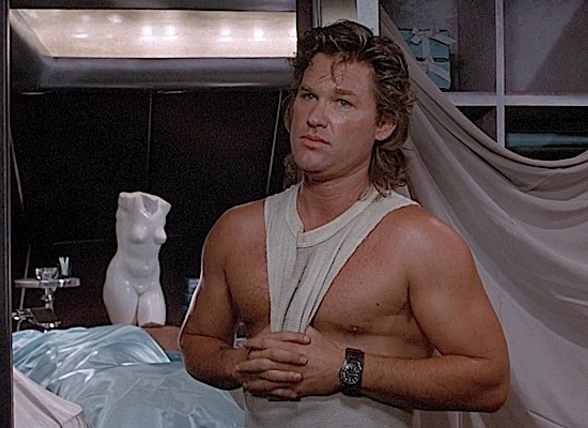 a shriekingly hot Kurt Russell in a tank top with goofy nudie statuary behind him