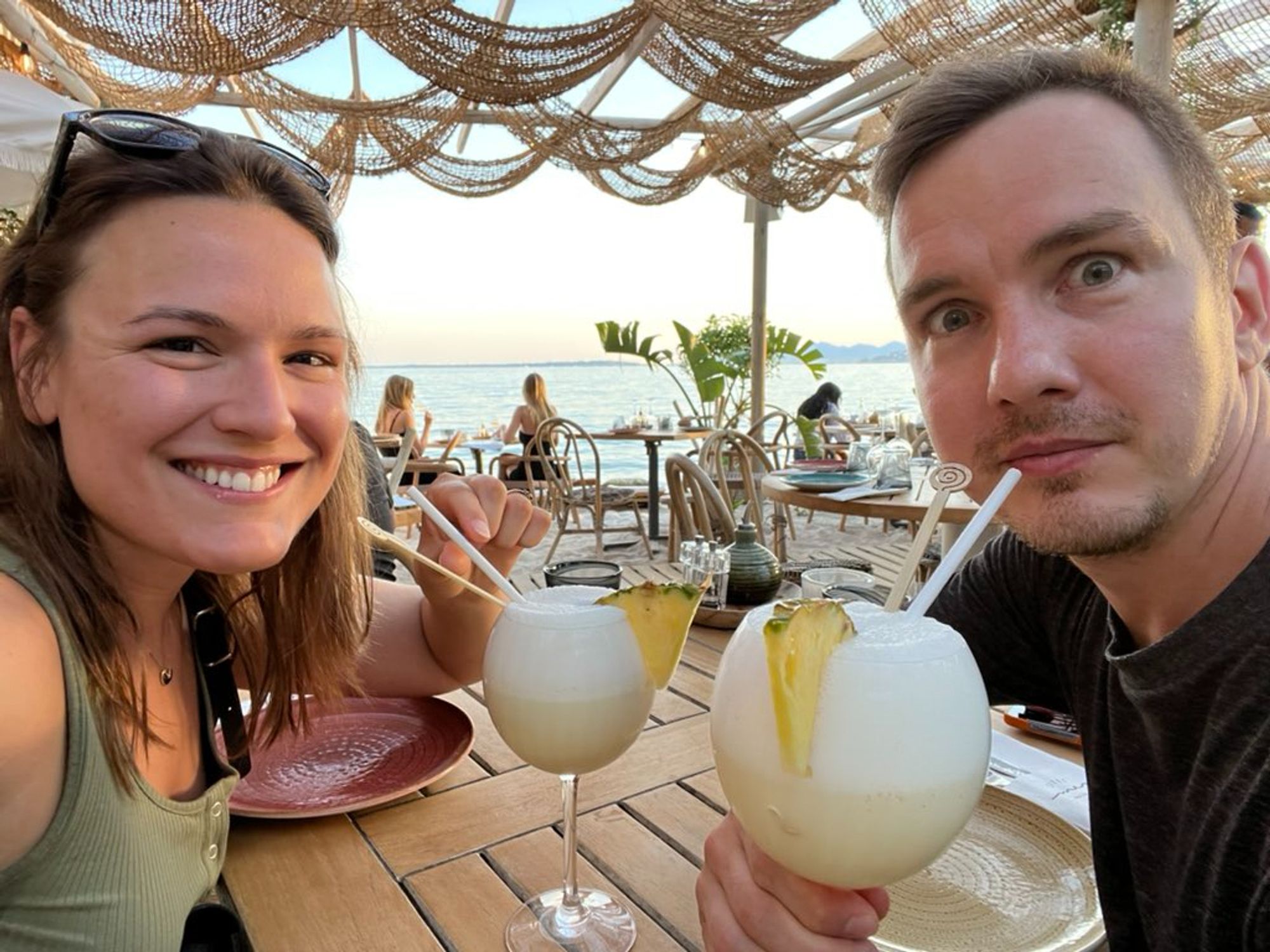 Oleg and Danica sipping piña coladas at a restaurant on the beach.