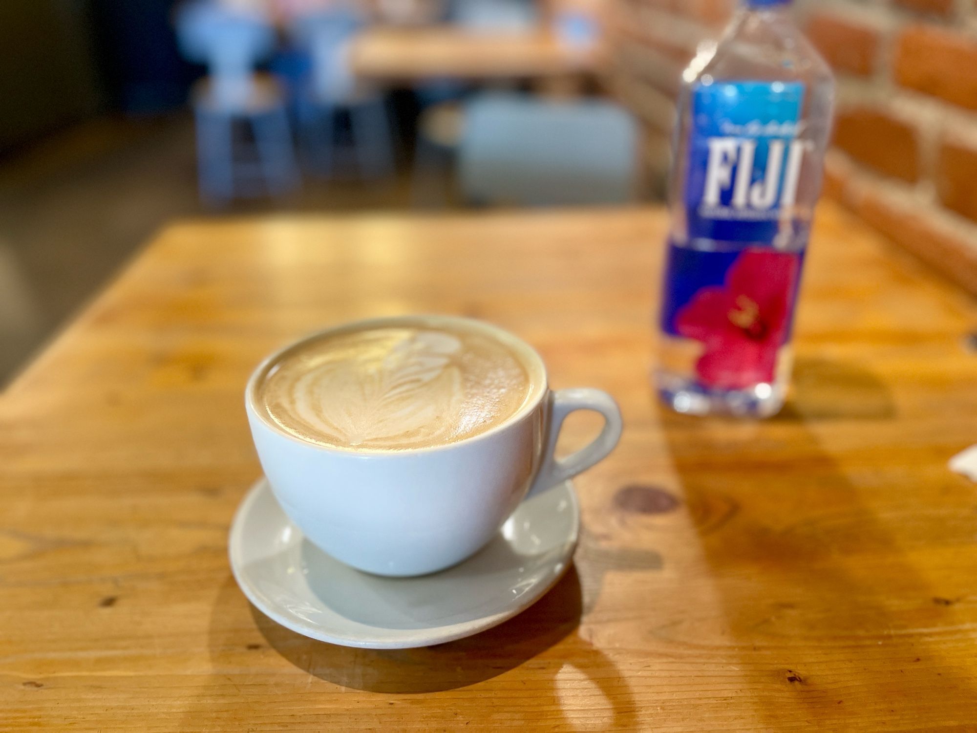 A photo of a white, wide rimmed, white coffee cup and saucer, filled with cappuccino and a bottle of Fiji water (blurred) to its right in the back.