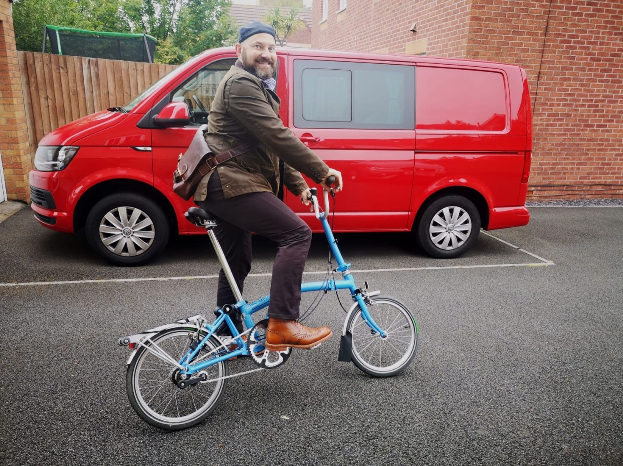 A middle-aged bearded man in a green Barbour jacket, dark grey troos and brown boots using a sky blue Brompton folding bicycle for his journey to work. No PPE or special clothing is in evidence.