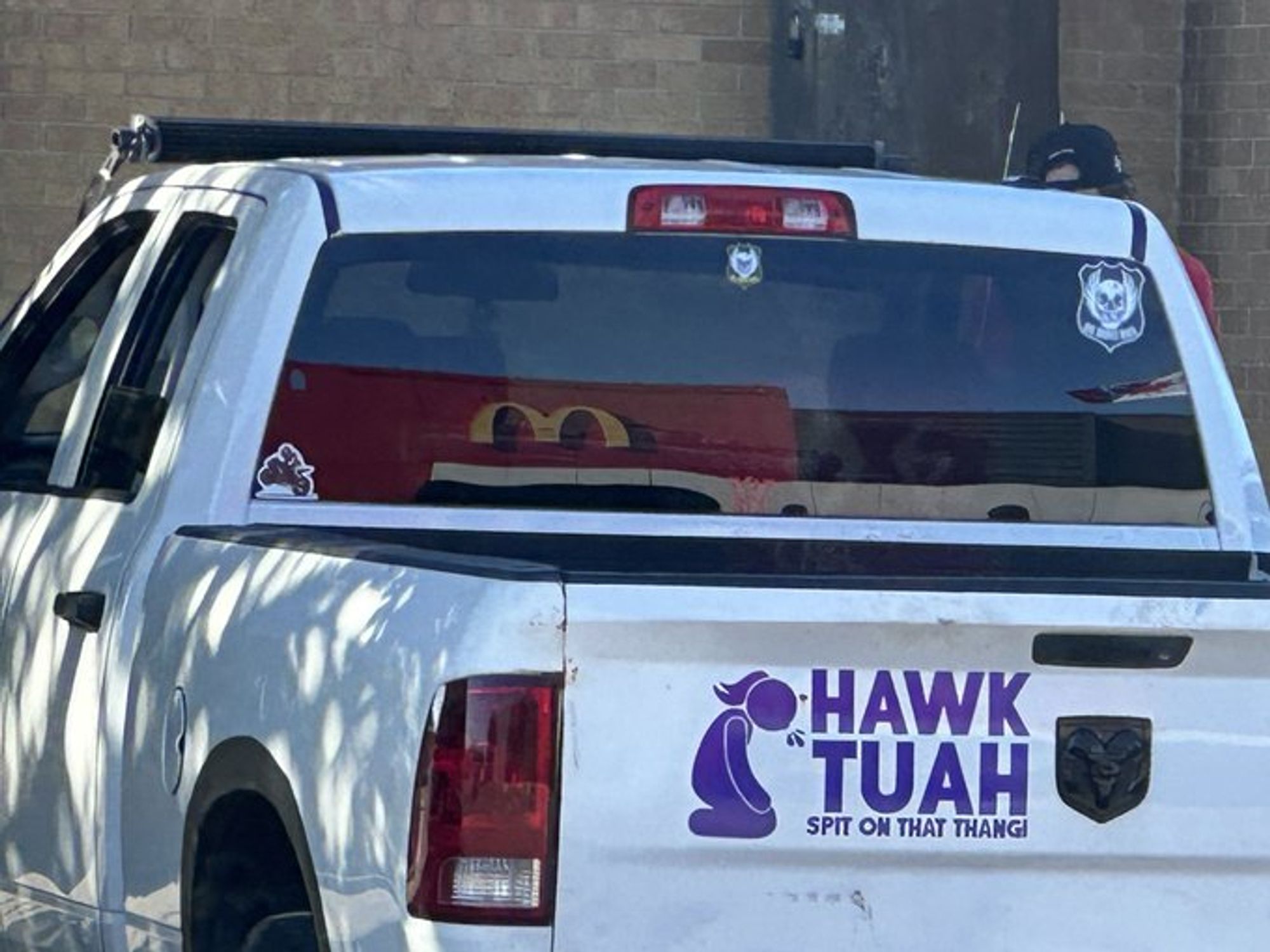 a stupid white pickup truck with a decal on the back showing a stick-figure woman kneeling and spitting, and the text "Hawk tuah spit on that thang!"