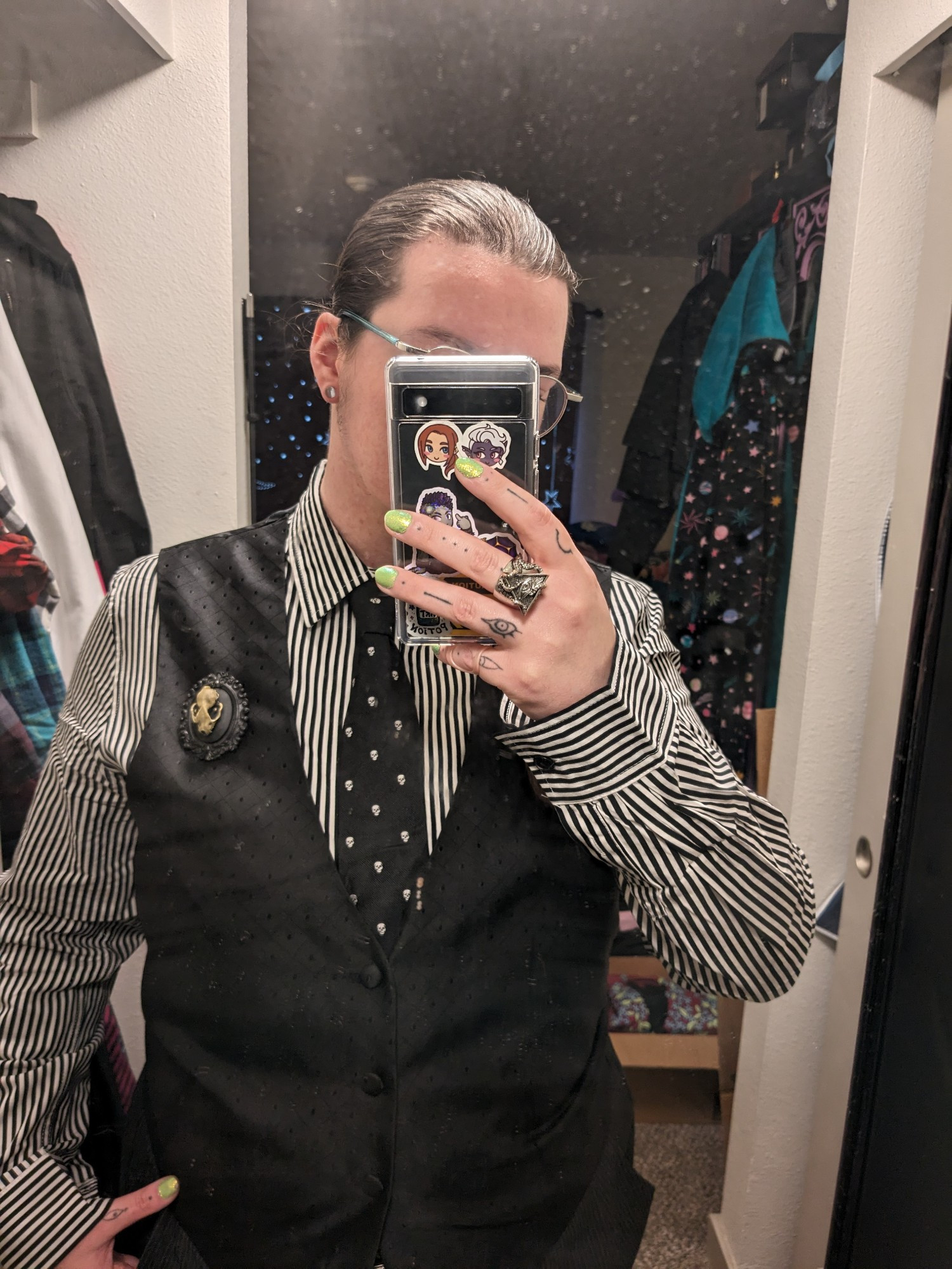 a close up mirror selfie of mochi in goth business wear from the waist up, clearly showing the narrow black and white stripe of their dress shirt, the bat skull of their brooch, and the skull dot pattern of their black tie, as well as the subtle black on black pattern of their waist coat. mochi has stylized eyes tattooed on their fingers and lime green nail polish