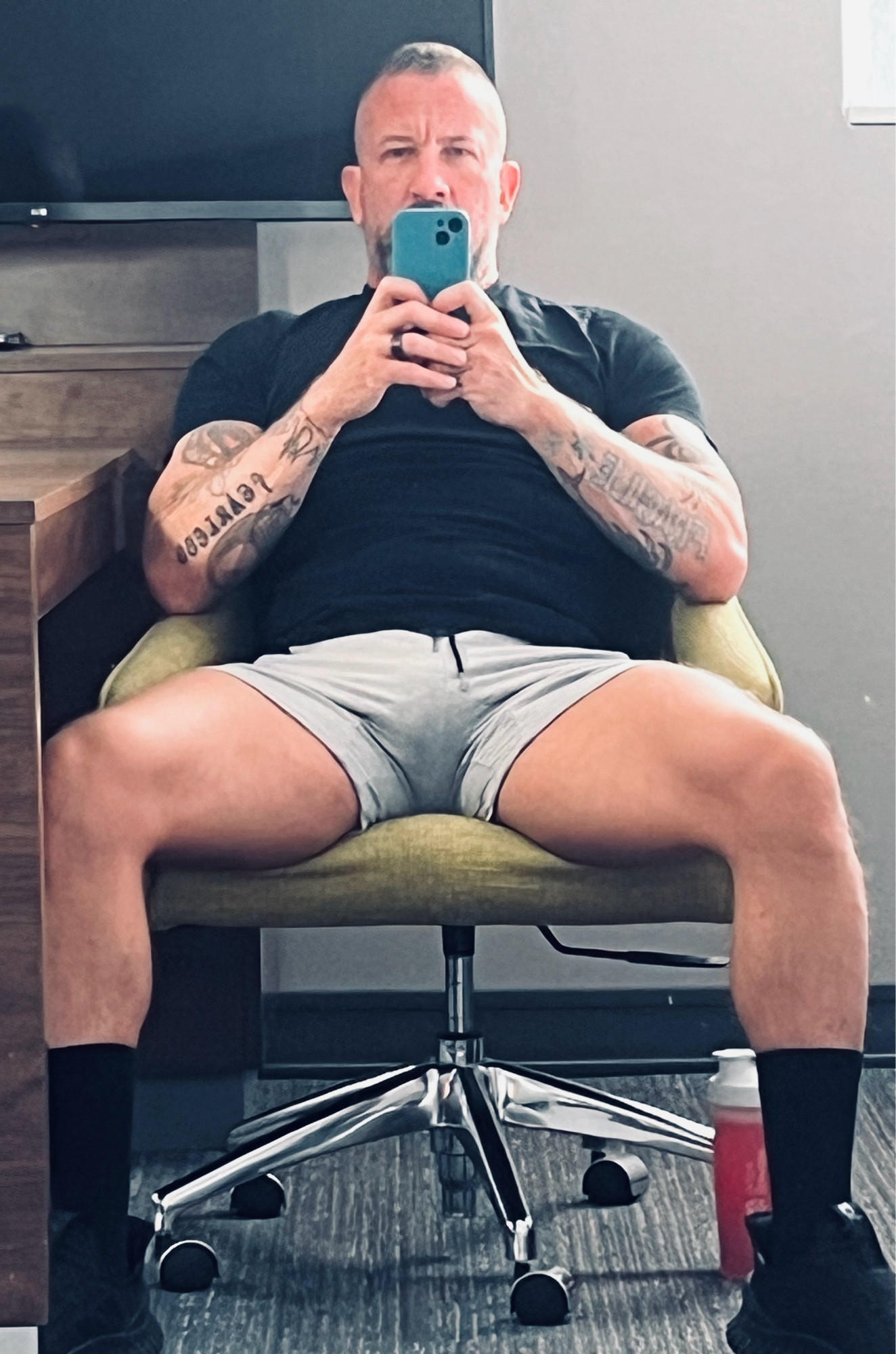 Coach is sitting in a wide-legged “manspread”, showing off his thick thighs. He’s dressed in a black tee and mid-calf socks, and pale gray shorts.