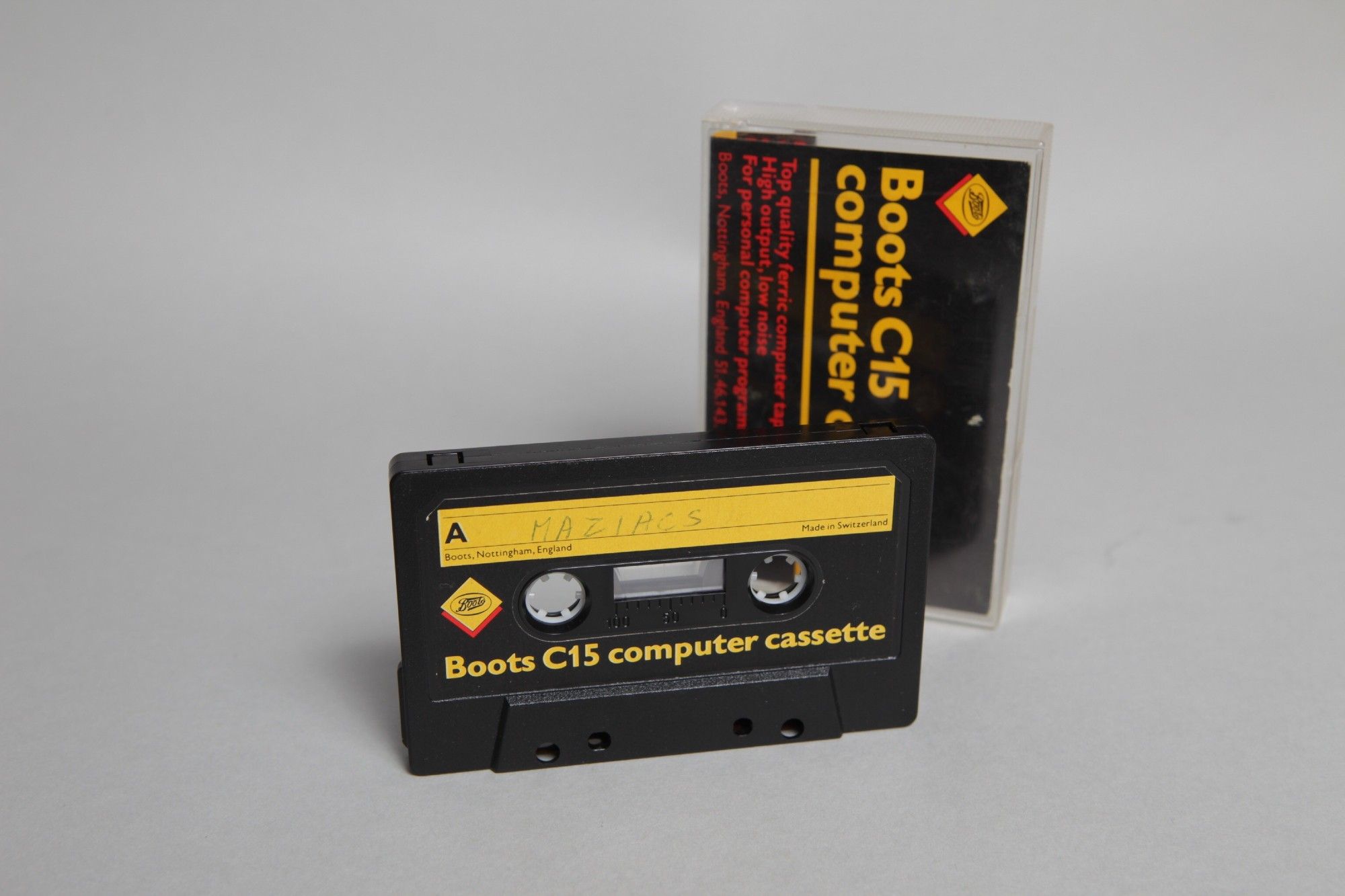 A Boots C15 cassette tape "backup" of the ZX Spectrum Maziacs game, on a grey background. In the foreground is the tape itself, with the box in the background. Both make use of a yellow and black colour scheme with red as a highlight.