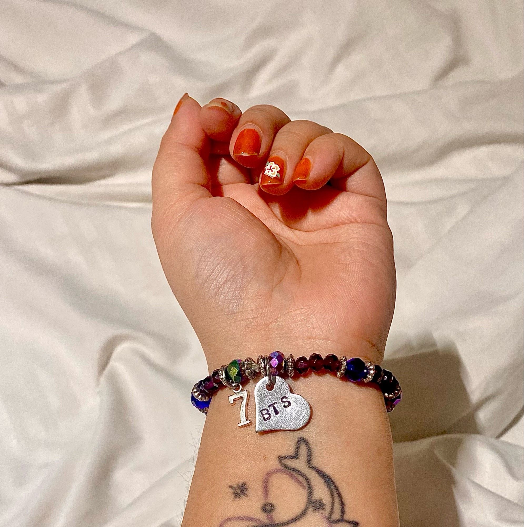 Picture, that I took, of my left hand with a BTS bracelet on. The bracelet has various crystal beads, mostly in different hues of purple and has a number 7 charm and a heart charm with ‘BTS’ engraved on it.