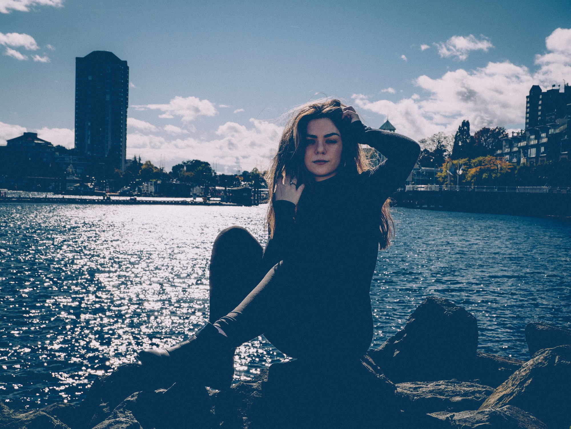 A woman with long dark hair sits on rocks in Nanaimo harbour wearing a black turtleneck, black pants and black boots.