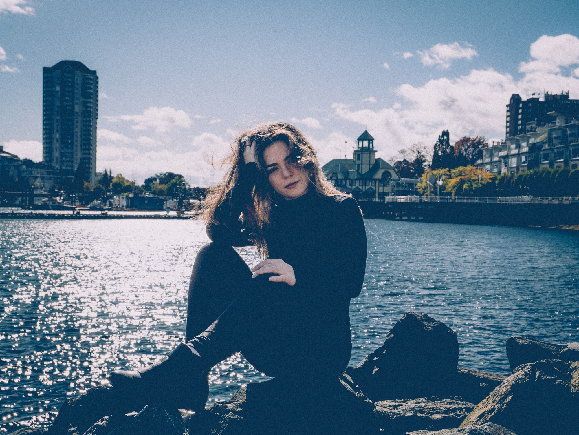 A woman with long dark hair sits on rocks in Nanaimo harbour wearing a black turtleneck, black pants and black boots.