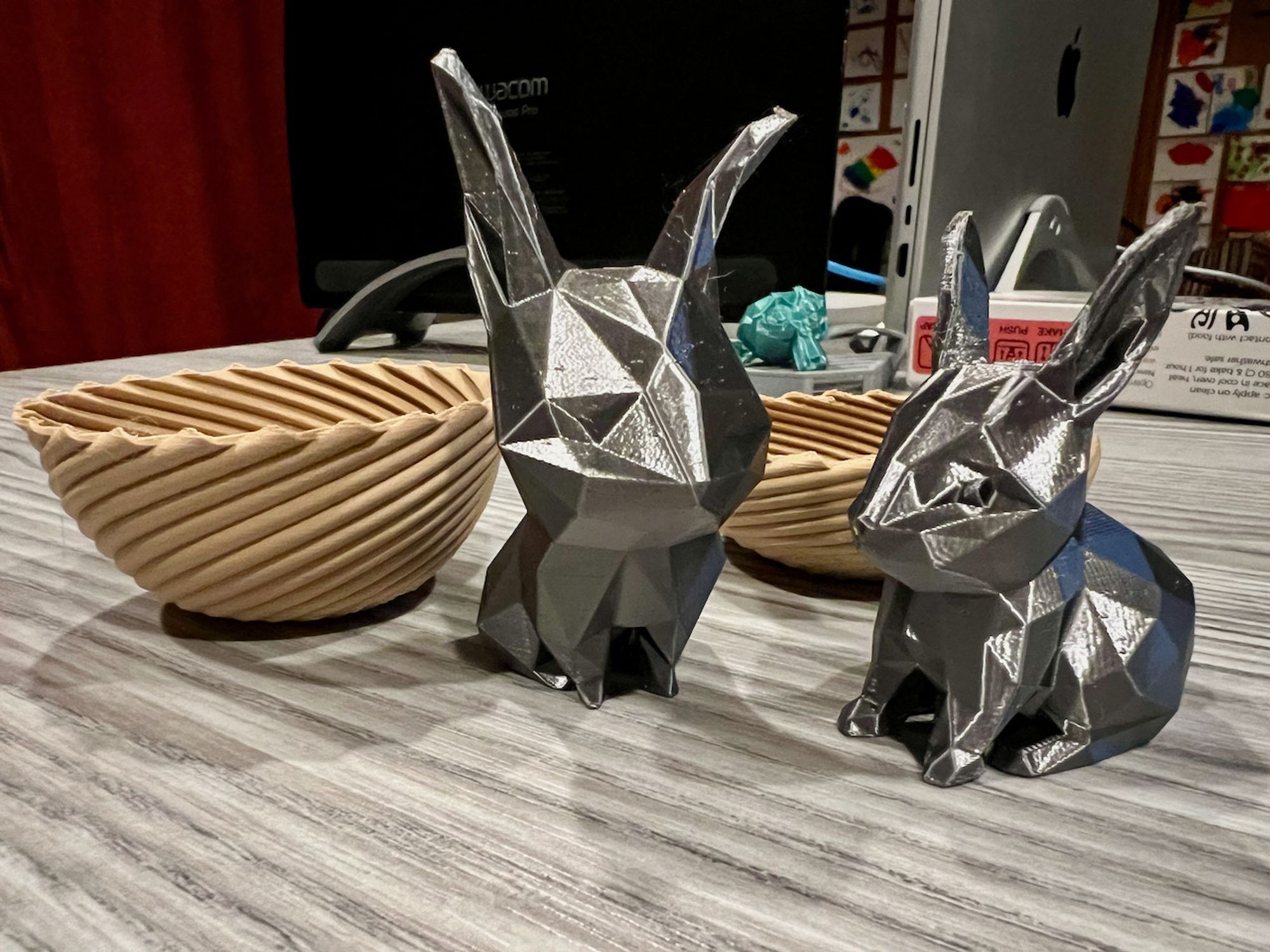 A photo of 2 printed tan bowls with a swirl pattern and 2 shiny silver low poly bunnies -- one with traditional proportions and another with a huge head, little feet, and tilted ears.