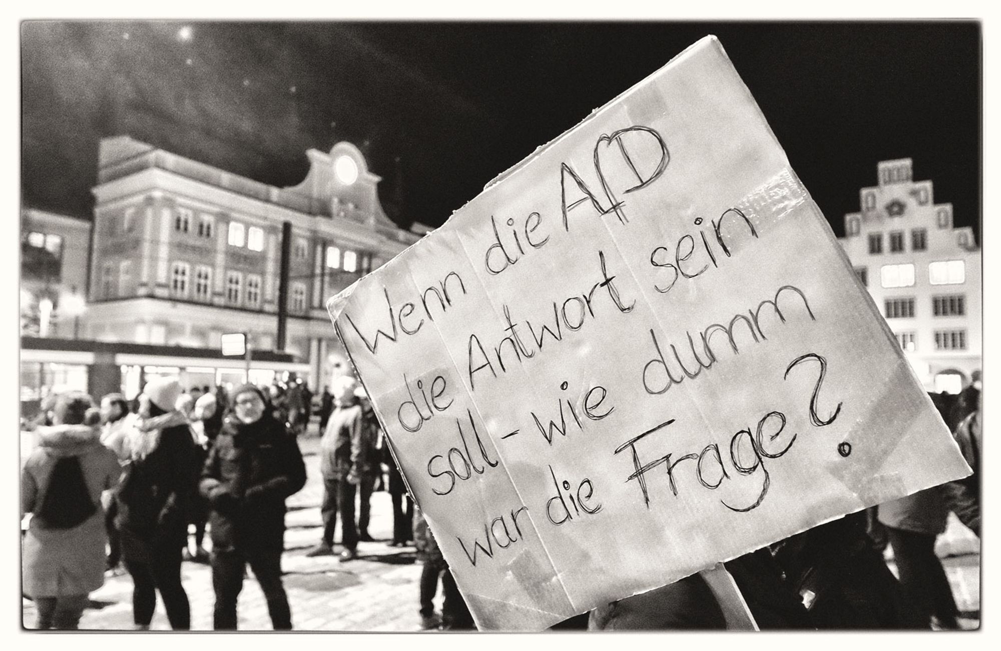 Foto von einer abendlichen Demonstration, auf dem Plakat im Vordergrund steht: „Wenn die AfD die Antwort sein soll - wie dumm war die Frage?”.