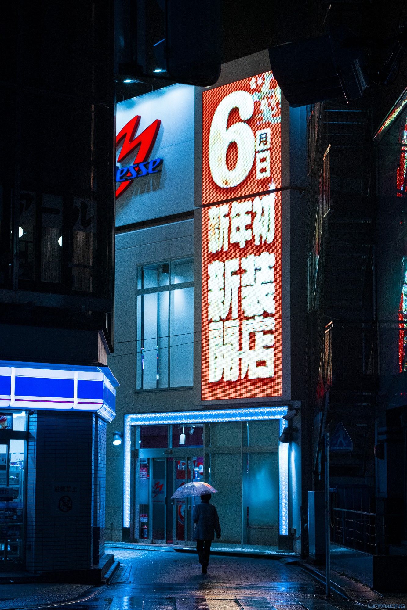 Photography by Liam Wong of Tokyo at night. A woman walks through the center of a tall vertical frame in the rain holding an umbrella. Overhead is large LED sign.