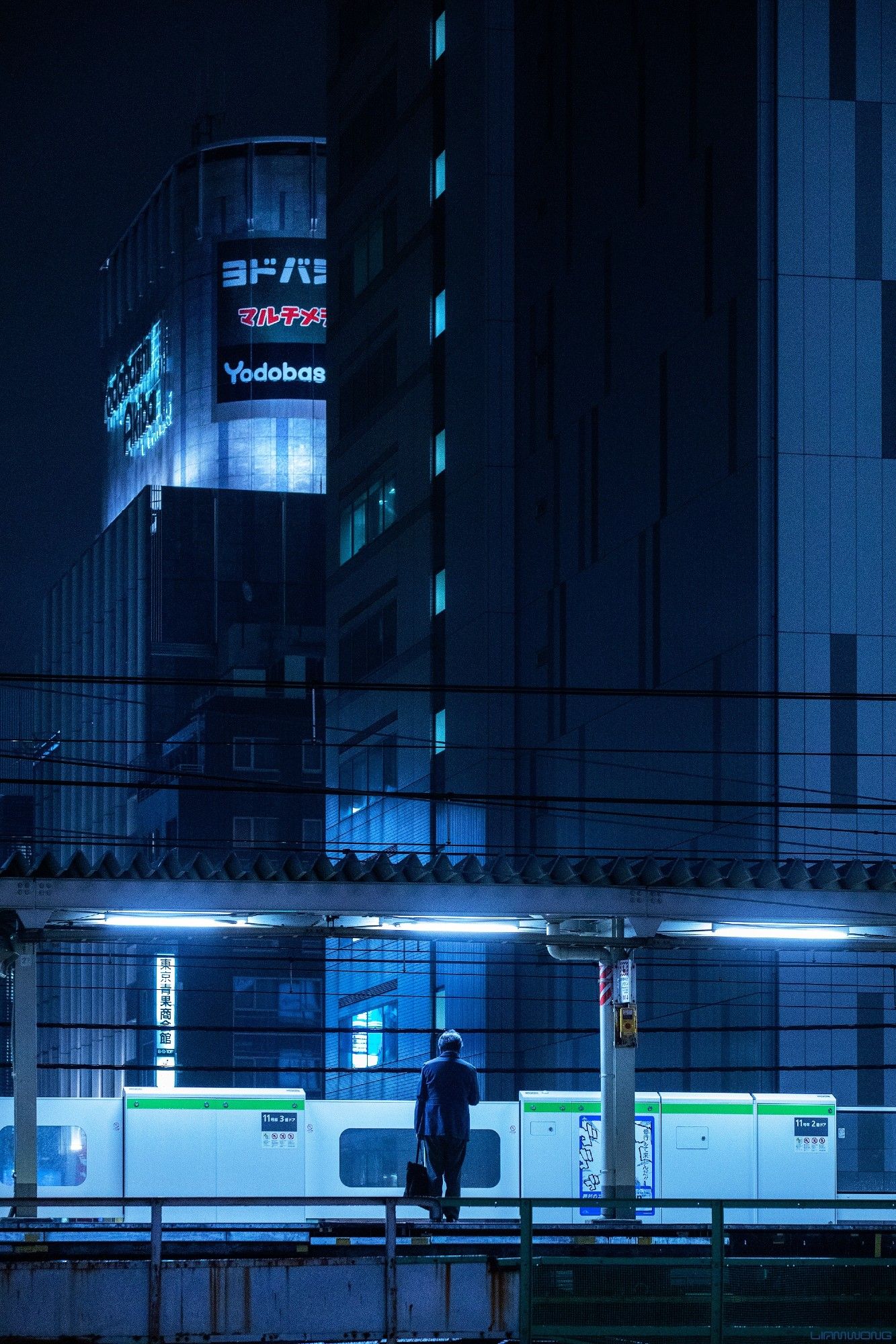 A businessman (salaryman) stands on an empty platform waiting for the last train home, in Tokyo Japan. The image is vertical with a cold blue sterile feeling to it. Above him and in the distance are large modern buildings, one sign can be seen briefly - YODOBASHI - one of the worlds largest electronic stores. He holds a briefcase and is lit up by halogen lights overhead. Overhead wires and buildings tower over him, he makes up a tenth of the image in scale. At first glance it may look like an illustration due to the colour treatment.