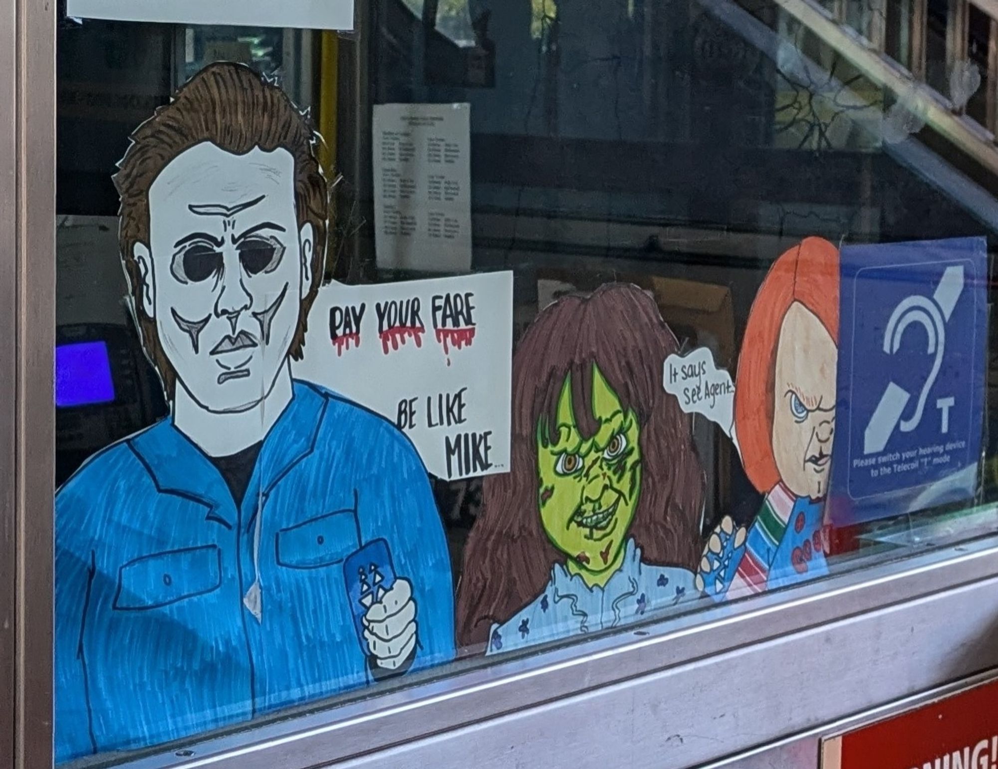 Transit station attendant's window. Three pieces of homemade art have been affixed to the glass with three horror movies characters. Left to right: the zombie-esque Michael Myers from Halloween, brown hair, blue jumpsuit, brandishing a Clipper card; a green skinned brown haired little girl I don't recognize (sue me), and the orange haired doll character Chucky from Child's Play.
Behind Myers is a sign: "Pay Your Fare - Be Like Mike" (first three words dripping blood)
Chucky has word balloon: "It says Pay Agent"