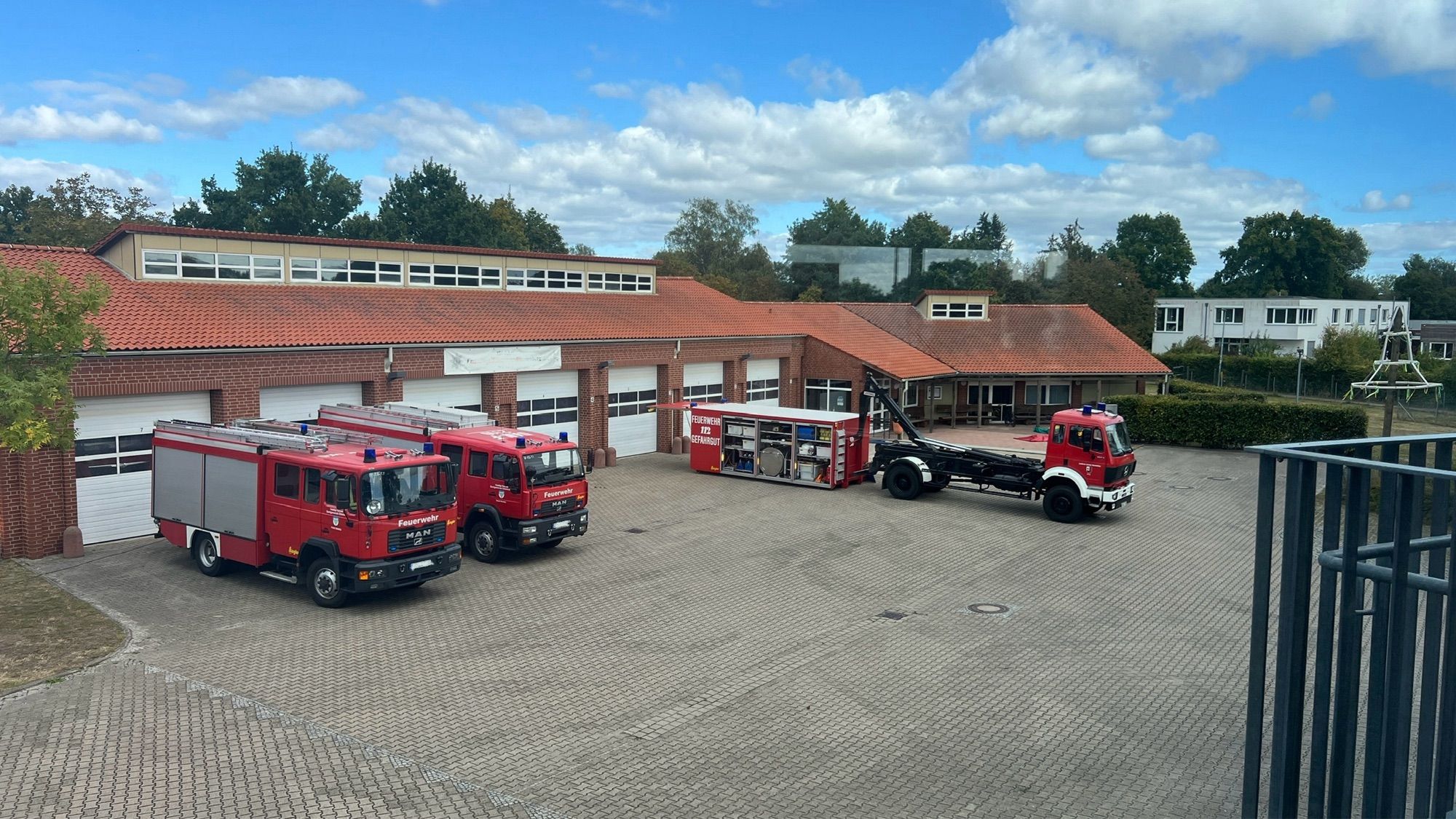 Das Bild zeigt eine Feuerwache mit mehreren Feuerwehrfahrzeugen, die vor den Garagen des Gebäudes geparkt sind. Drei rote Feuerwehrfahrzeuge, darunter ein MAN-Feuerwehrauto, stehen auf einem gepflasterten Hof. Ein weiteres Fahrzeug mit einem Abrollbehälter, auf dem “Feuerwehr für Gefahrgut” steht, ist ebenfalls zu sehen. Das Gebäude hat ein rotes Ziegeldach und mehrere Garagentore. Im Hintergrund befinden sich Bäume und einige Gebäude, der Himmel ist teilweise bewölkt, aber größtenteils blau.