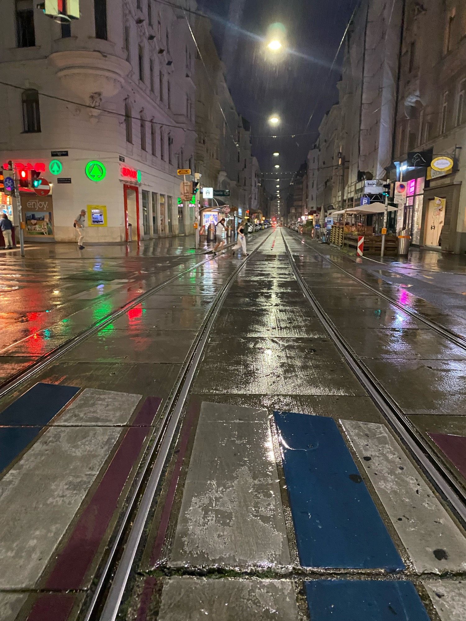 Regennasse Straße bei Nacht in Wien, im Vordergrund ein Zebrastreifen. Die Lichter der Straßenbeleuchtung und Geschäfte spiegeln sich auf der nassen Fahrbahn. Straßenbahnschienen führen umsäumt von drei- bis vierstöckigen Häusern in die Bildmitte