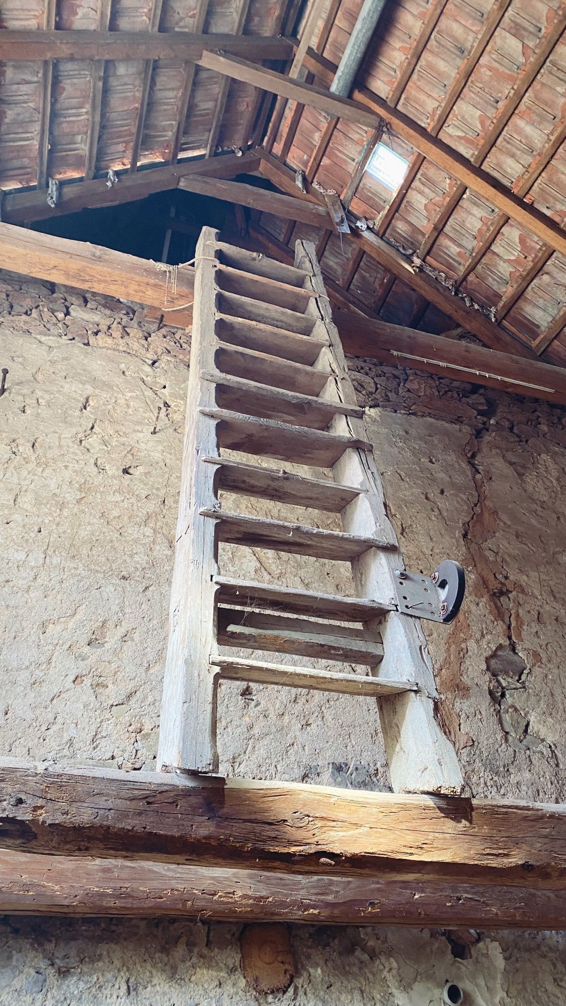 Eine große Holzleiter steht vor einer rissigen Mauer auf einem grob behauenen Querbalken und führt so unters Dach, das man gerade noch von unten sieht