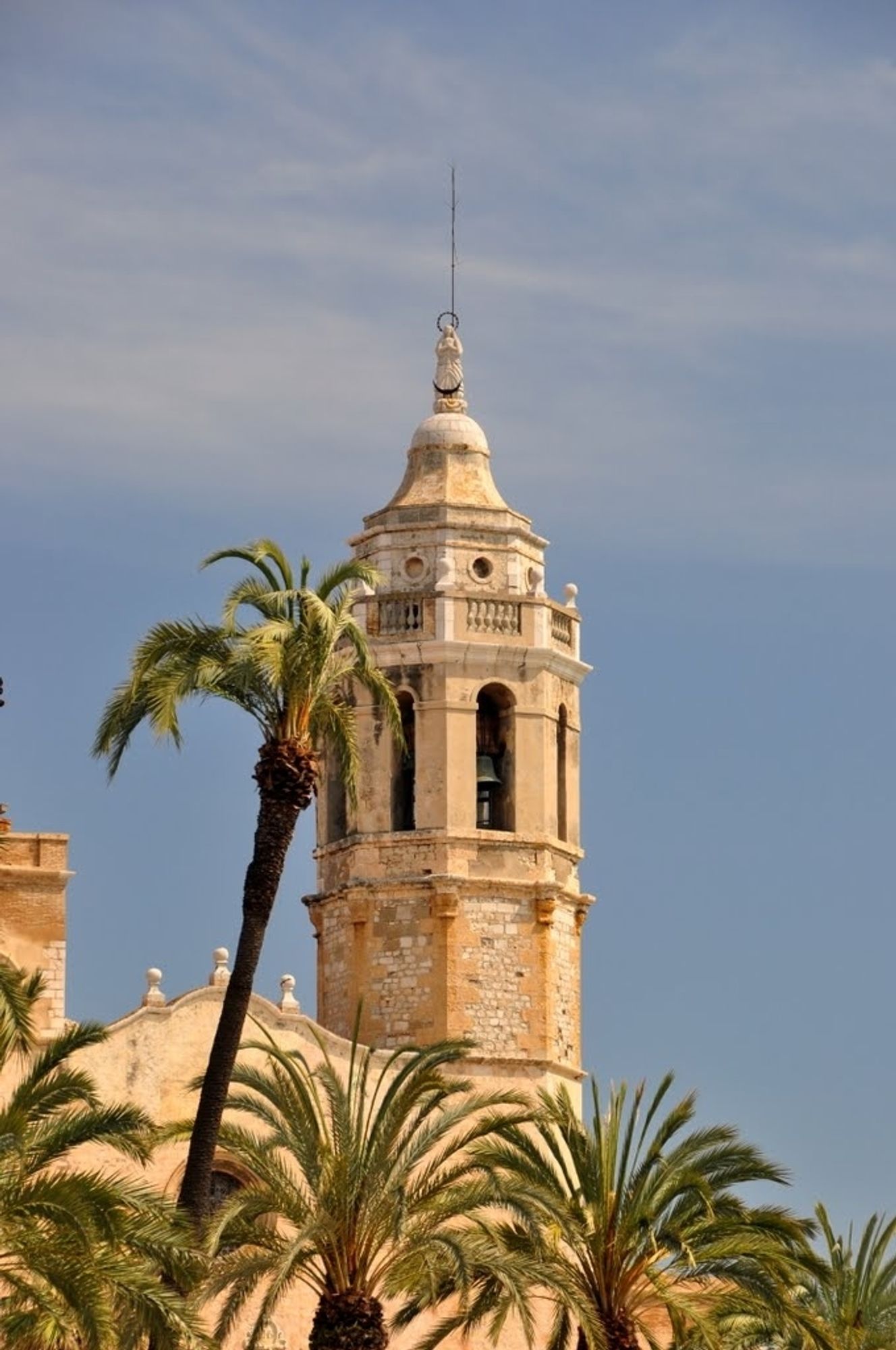 Ein mediterraner  Kirchturm in gelb und etwas verwittert. Im unteren Viertel ist eine Reihe Palmenkronen zu sehen. Eine weitere Palme ist mit leicht nach rechts geneigtem Stemm dicht links neben dem Kirchturm zu sehen.