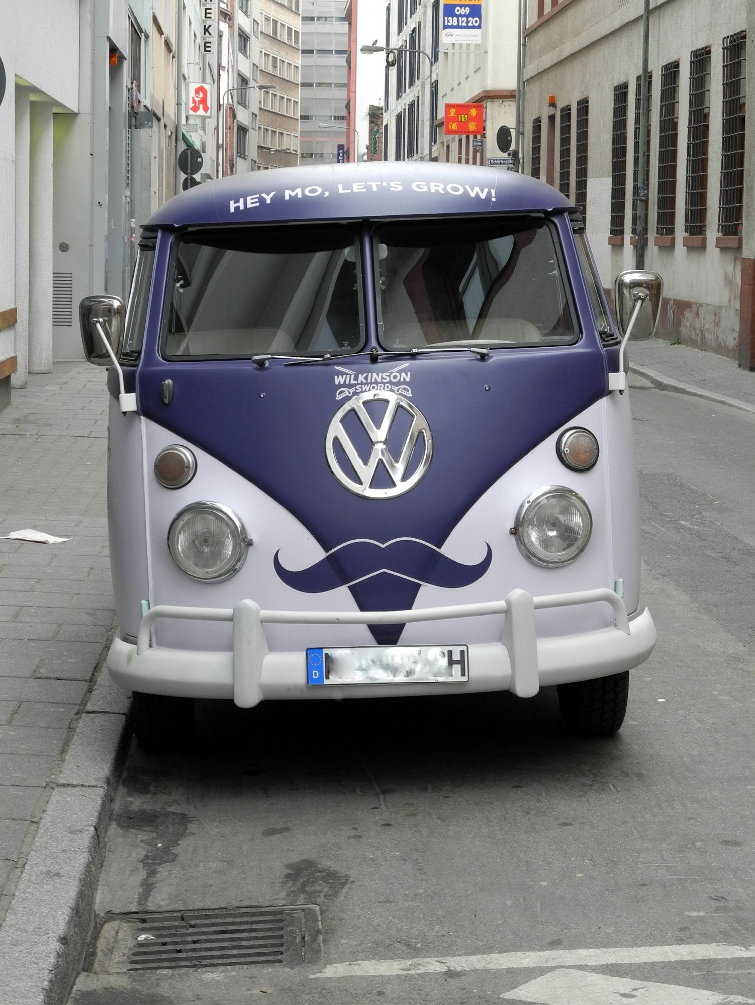 Hochformat. Ein VW Bus älteren Datums von vorne. Der Bus ist Dunkellila Helllila lackiert. Unter den der geteilten Windschutzscheibe ist groß, rund und silbern das VW-Logo zu sehen. Links und rechts davon in orange je ein runder Blinker und darunter in weiß je rechts und links ein Scheinwerfer. Zwischen den beiden runden weißen Schinwerfern ist ein Schnurrbart in dunkellila zu sehen. Über der Windschutzscheibe steht: HEY MO, LET'S GROW!