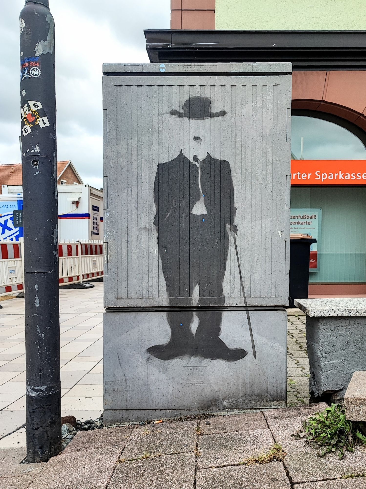 Hochformat. Auf einem grauen Stromkasten ist in Schwarz Charlie Chaplin zu sehen. Dabei sind nur die schwarzen Teile zu sehen, d.h. Hut, Schnurbart, Jacket, Stock und Schuhe.