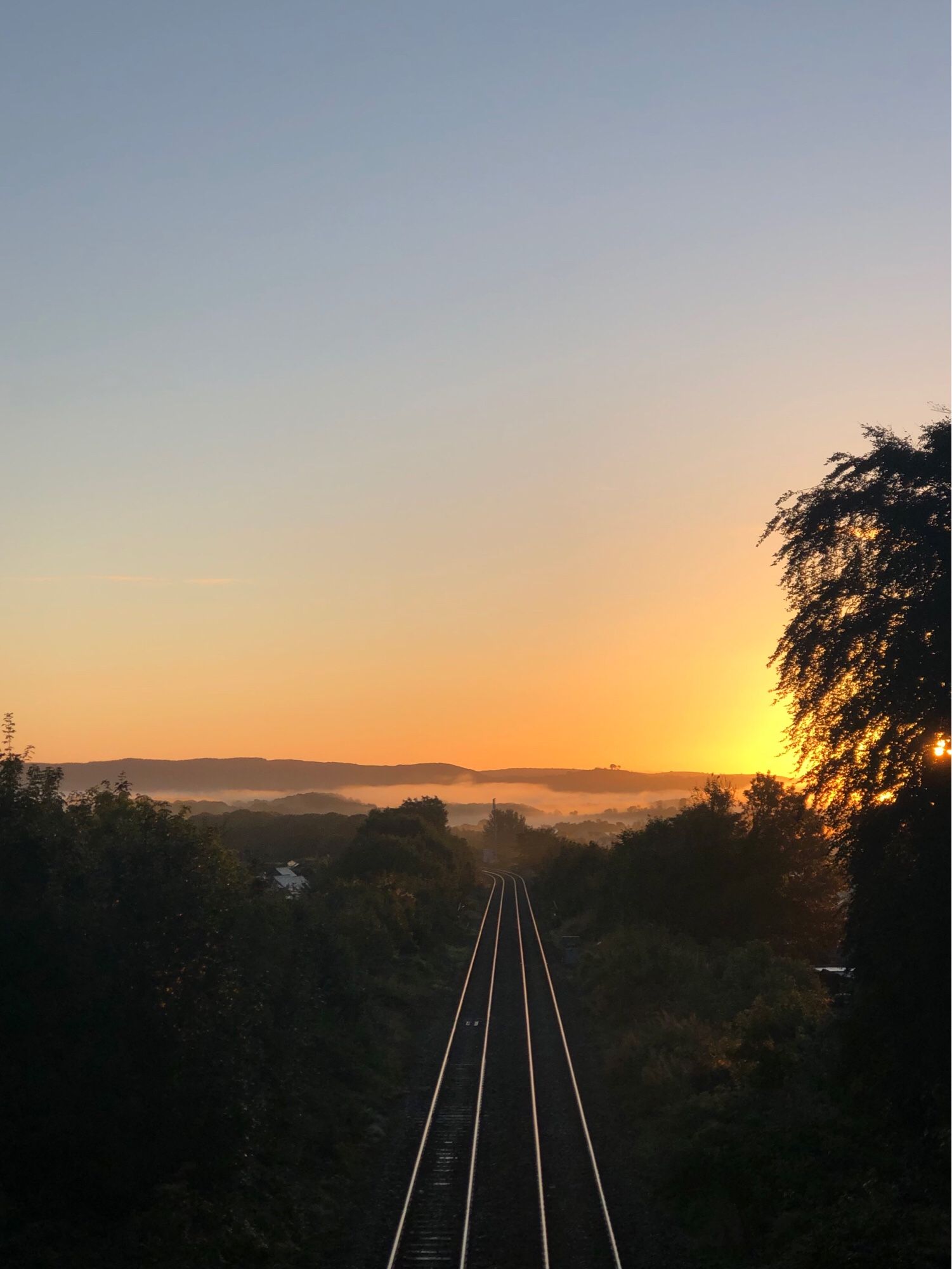 Train lines run vertically centre frame. The trees and shrubs on the banks either side are in darkness. There is low lying cloud in the distance and in front of a dark fell line. The sun is rising, hot yellow glow right frame behind a tree. The sky is beautiful pastel shades of orange and pink fading to palest blue higher up.