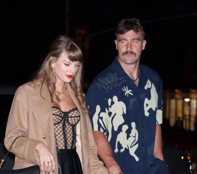 A photo of Taylor Swift and her hunk of the year, Travis Kelce, stepping out somewhere: she's in a tan overcoat with a Dior bustier underneath and he's dressed in a dark blue Hawaiian-style short-sleeve with a white print that may or may not be horses and people. He is wearing a moustache and a haircut closely resembling Robin Williams circa 1990.