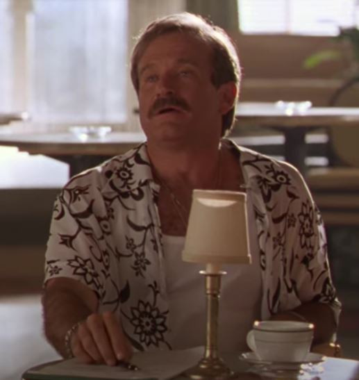 Screenshot of Robin Williams from The Birdcage (1995). He is seated at a small table, wearing a short sleeve shirt with flowery patterns and a tank top underneath. 