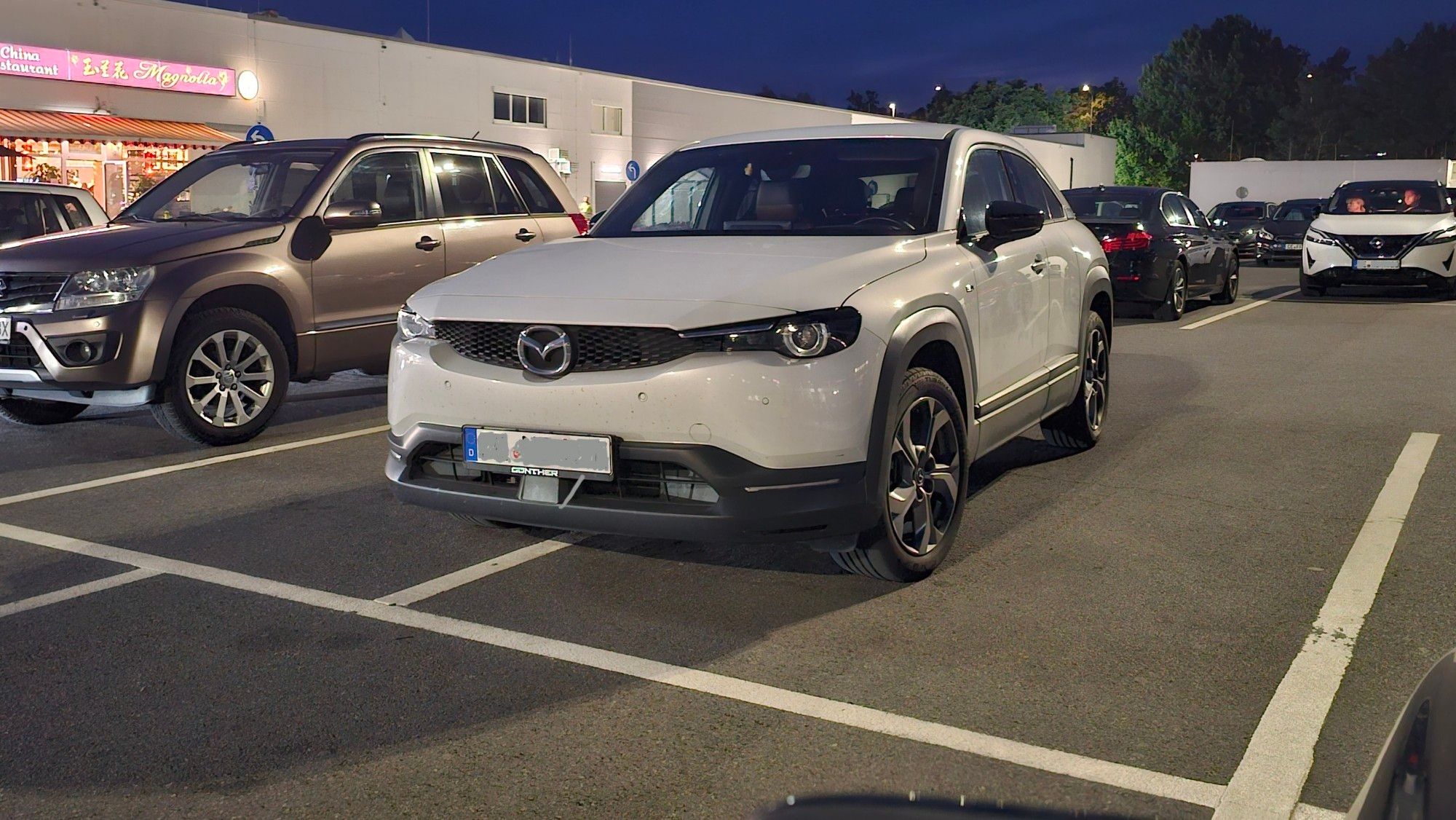 Weißer Matzda-SUV auf dem Parkplatz eines Einkaufszentrums. Das Foto ist gegen Abend aufgenommen, es dämmert und die Lichtmasten sorgen für die Beleuchtung.
Der fahrende Jupitermond hat sich genau in die Mitte von zwei Parkplätze gestellt.