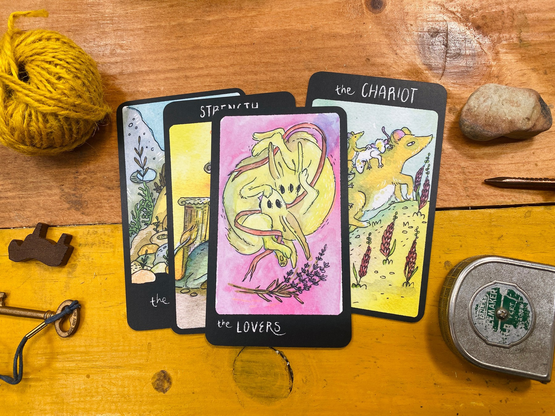 A close up of some tarot cards; the Lovers and the Chariot are seen - the Lovers are two intertwined three eyed rabbit-esque ceatures, and the Chariot is an exhausted possum.