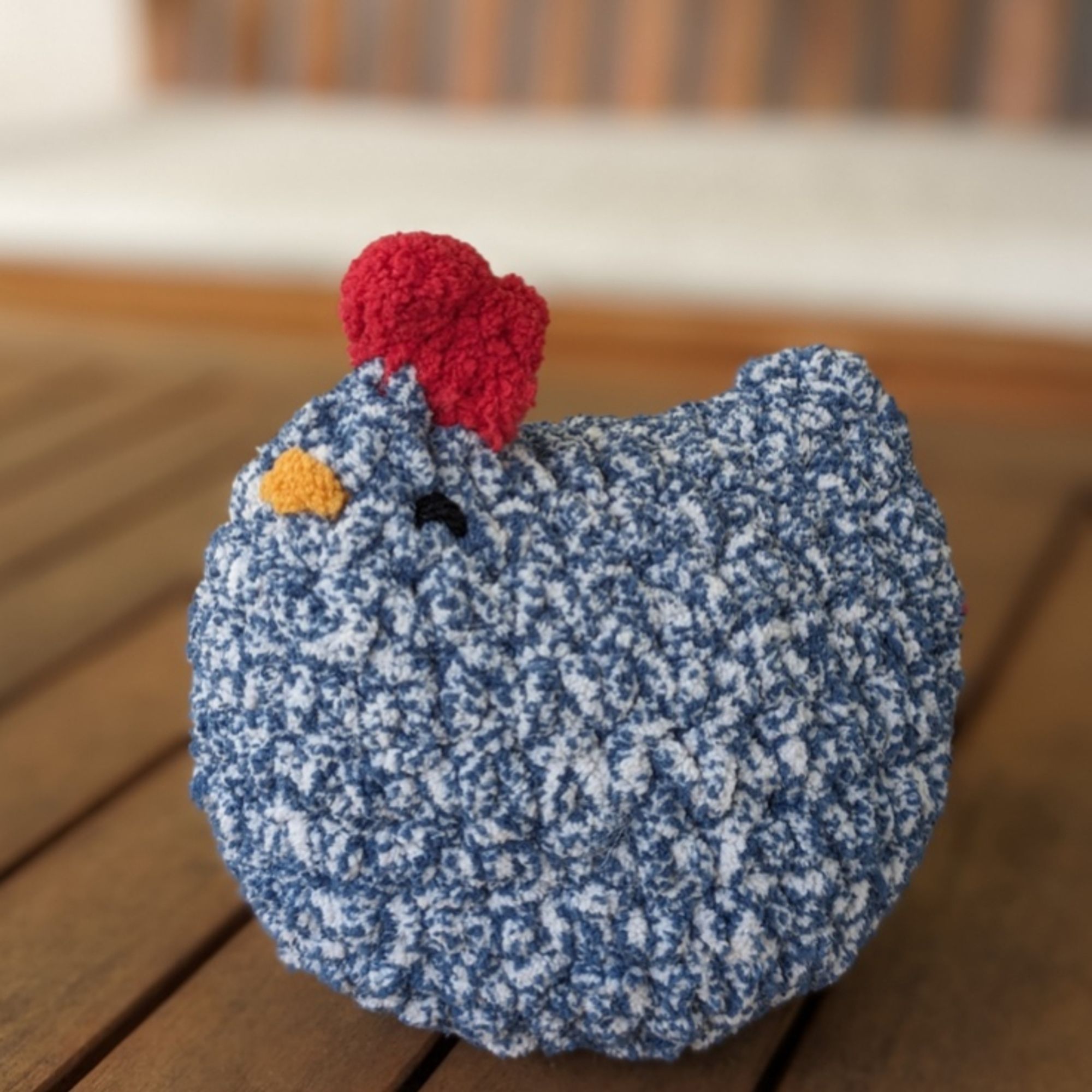 a photo of a medium sized, handmade, crochet chicken made from blue speckle, plush yarn with a small yellow mouth, embroidered black eyes that make it look like its smiling and a red comb. It is very round and appears quite squishable!