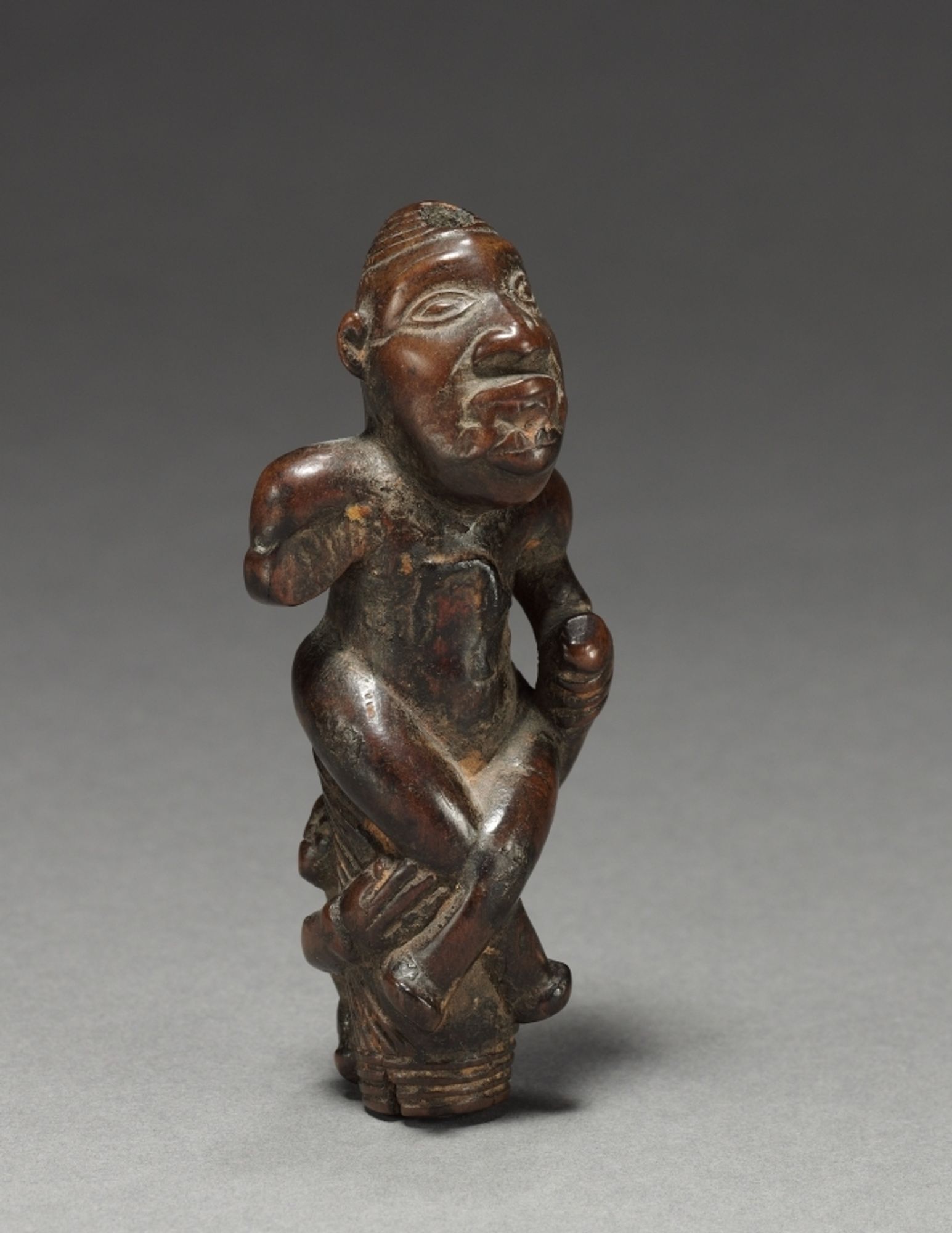 This male figurine was probably once attached to a scepter or perhaps even to a fly whisk. There are traces on the surface that indicate the sculpture may have carried a medicine-filled package on its abdomen. The man seated with legs crossed and wearing a coiffure of Portuguese inspiration may represent a chief. His broken-off right hand would have held the carved imitation of a root called <em>munkwiza</em>, one of the most important chiefly medicines often buried in the earth of a chief 's compound. Perhaps the smaller figurine on the rear of the work, beneath the root-chewing chief, depicts a child holding onto his parent's back, though such imagery is more typically seen on female sculptures.