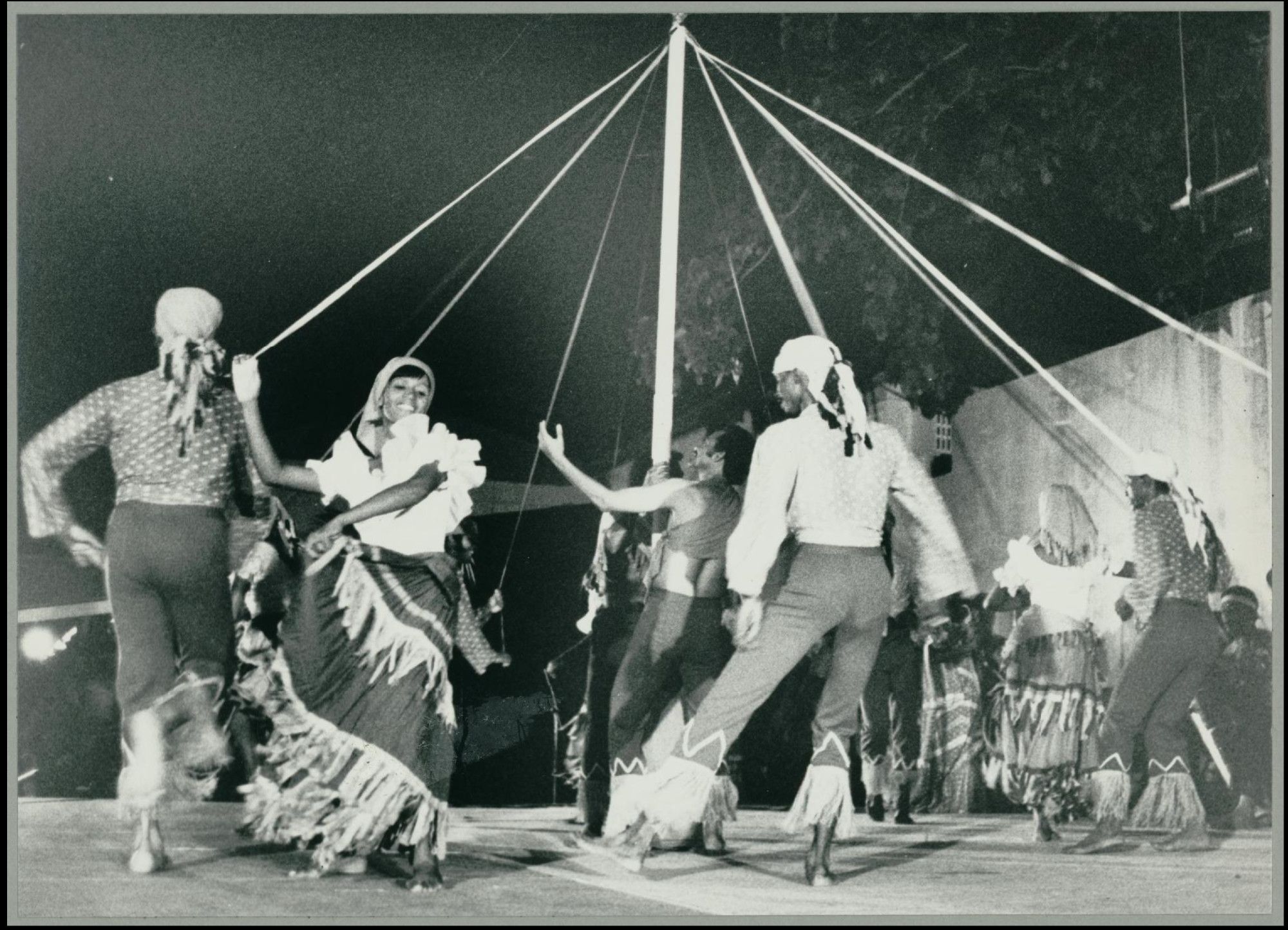 Black and white photograph of people from Barbados dancing in folk costumes around a maypole. "... participants dance in a circle, each holding a colored ribbon attached to a pole. The ribbons are intertwined and plaited either to the pole itself or into a web around the pole. The dancers then can retrace their steps in order to unravel the ribbons. "