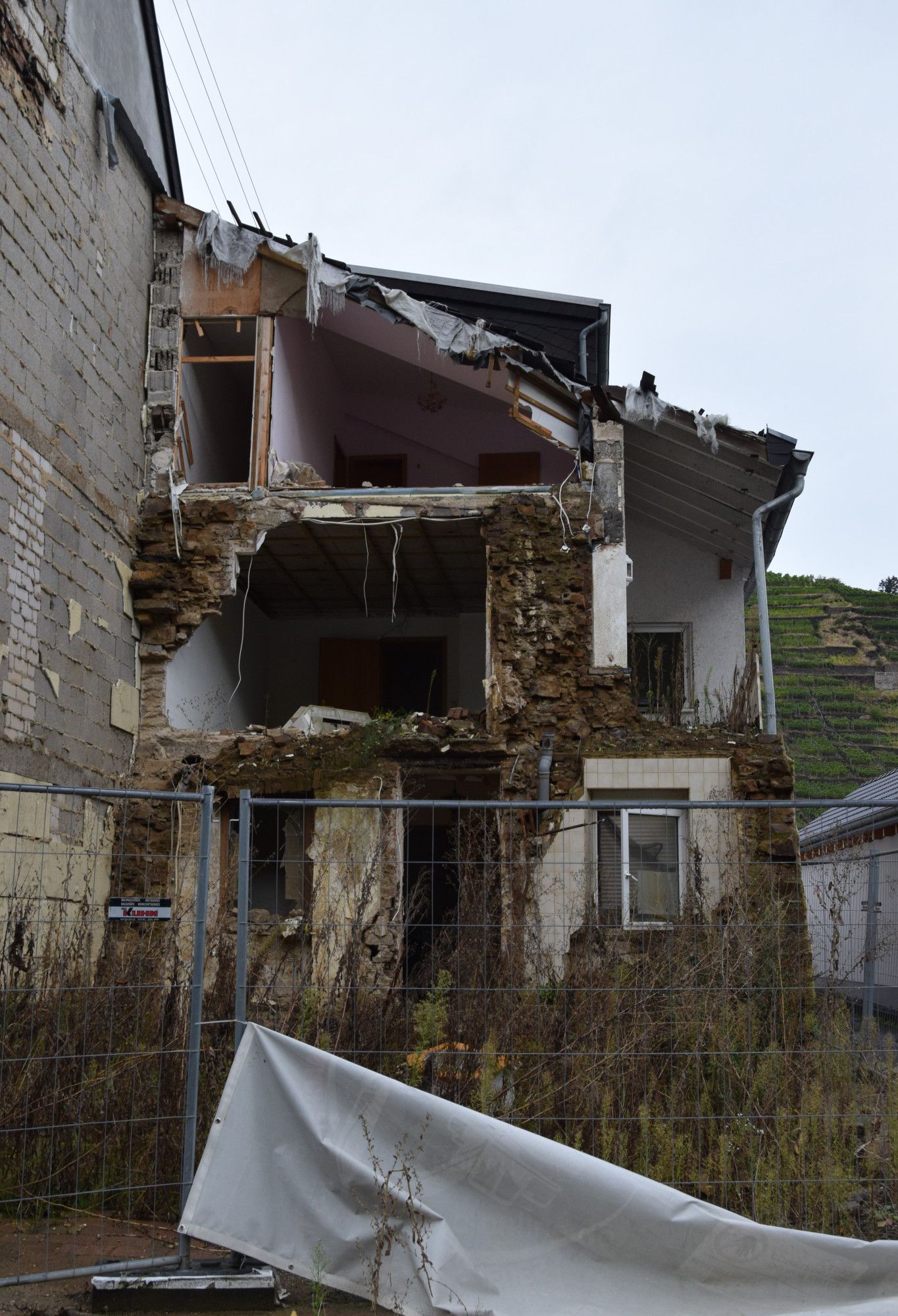 Eine ruiniertes Haus, die Vorderfront fehlt total, man blickt in offene Zimmer. Davor ein Bauzaun mit einer Plane.