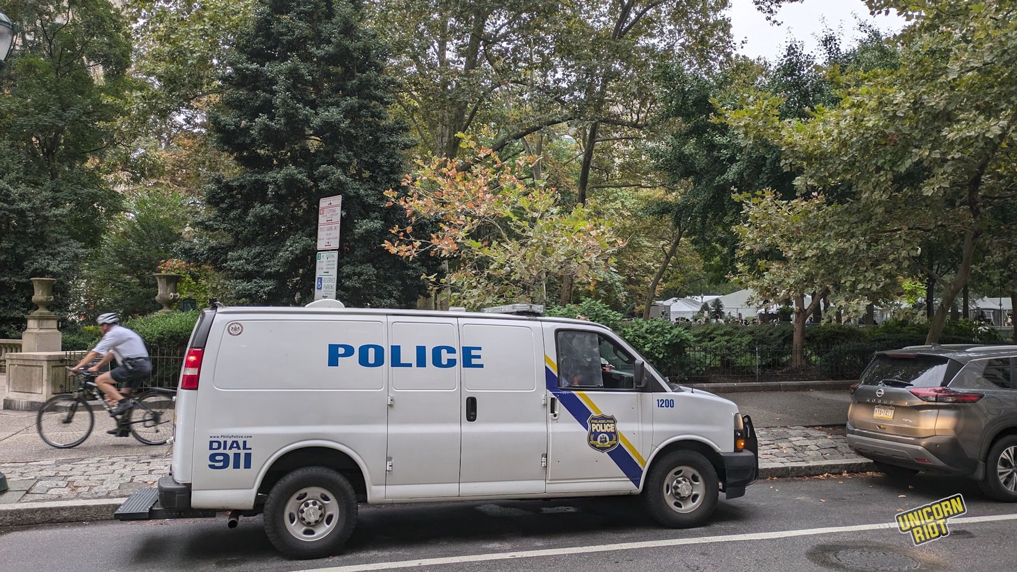 Same Philly Police van seen from across the street outside Rittenhouse as a bike cop bikes down the sidewalk by the park moving from right to left across our frame - almost out of the shot on the left side