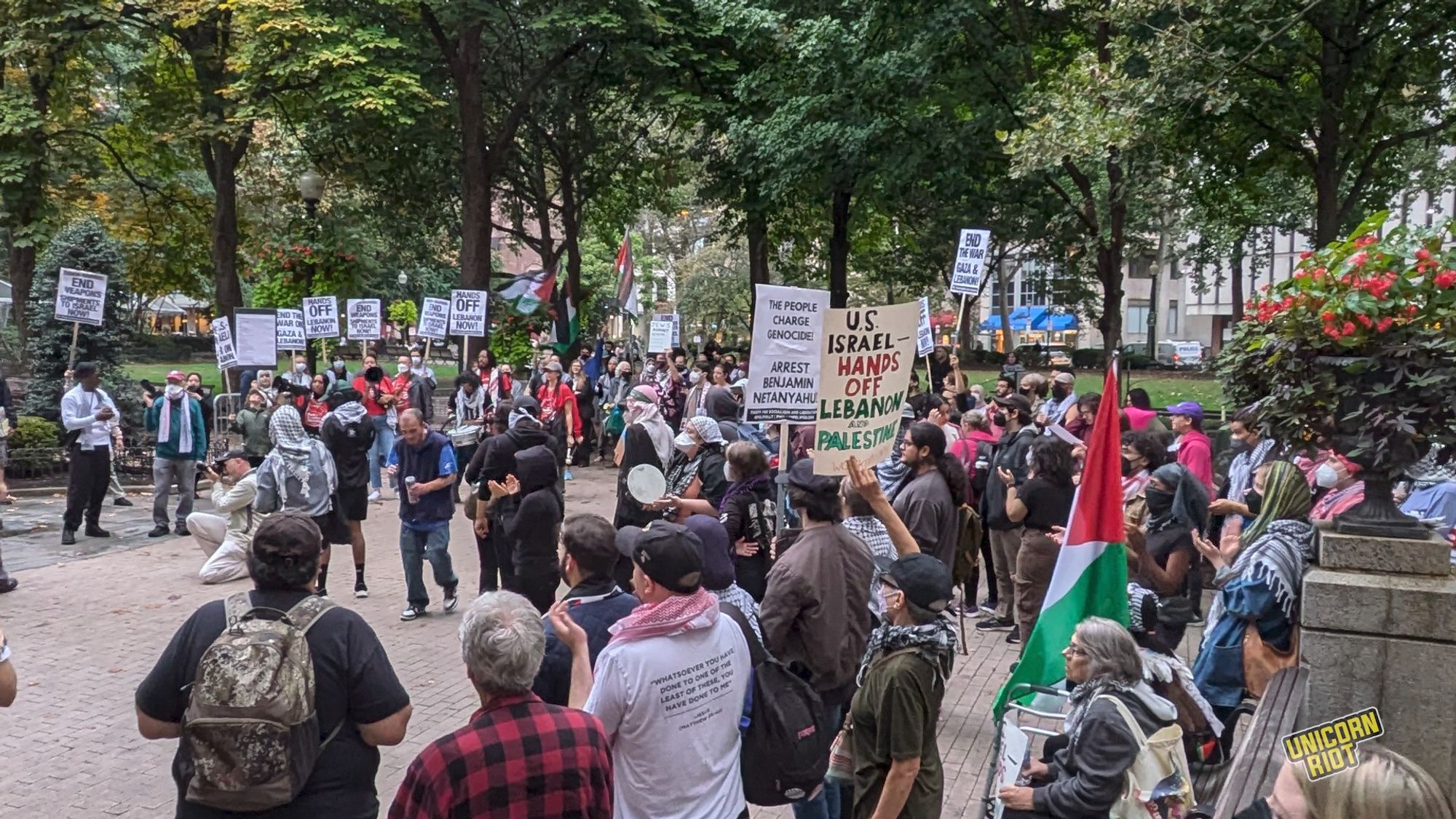 A group of protesters stands in a tree-ringed plaza holding Palestinian flags and protest signs with messages like "US Israel - Hands off Lebanon and Palestine"