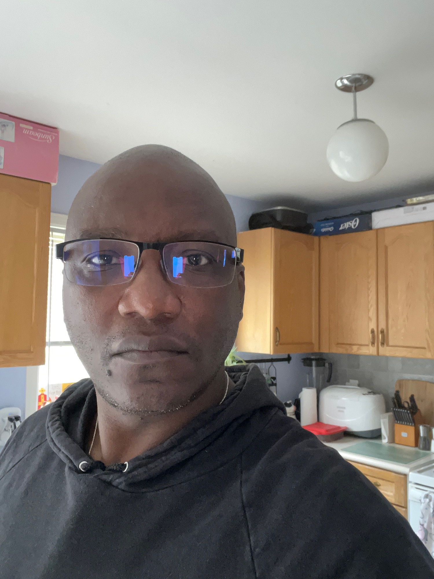 Head and shoulders photo of a bald black man wearing glasses. He’s standing in a kitchen. 