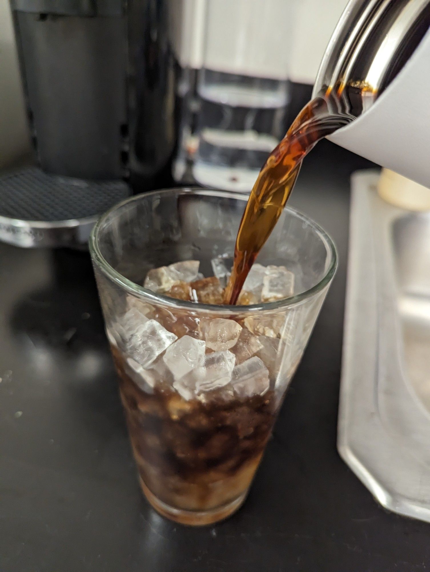 pint glass with ice pellets sitting on a black countertop, being filled with coffee from a thermos