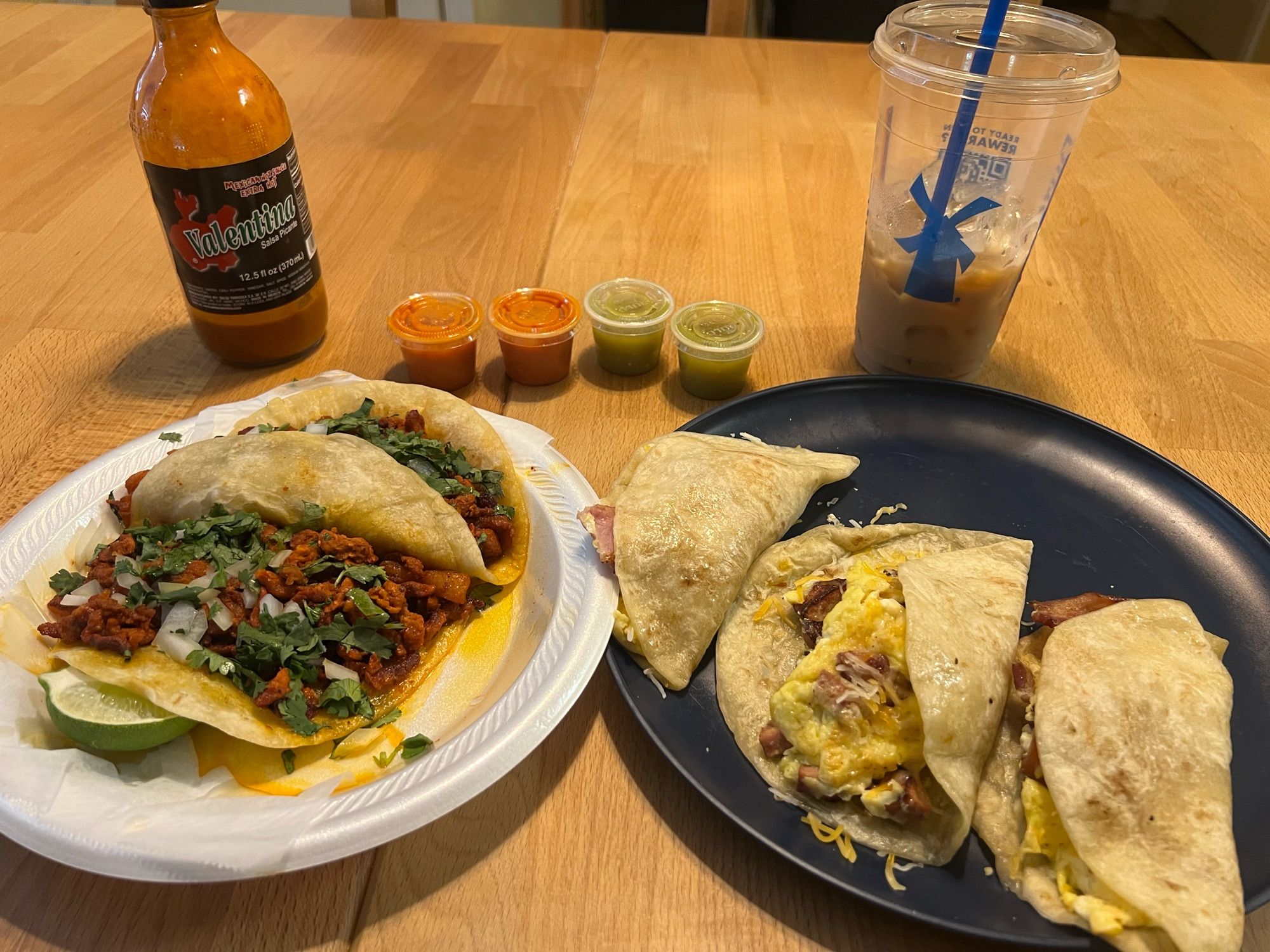 A spread of five tacos (two al pastor and three breakfast tacos) on a table by a hot sauce bottle and an iced coffee