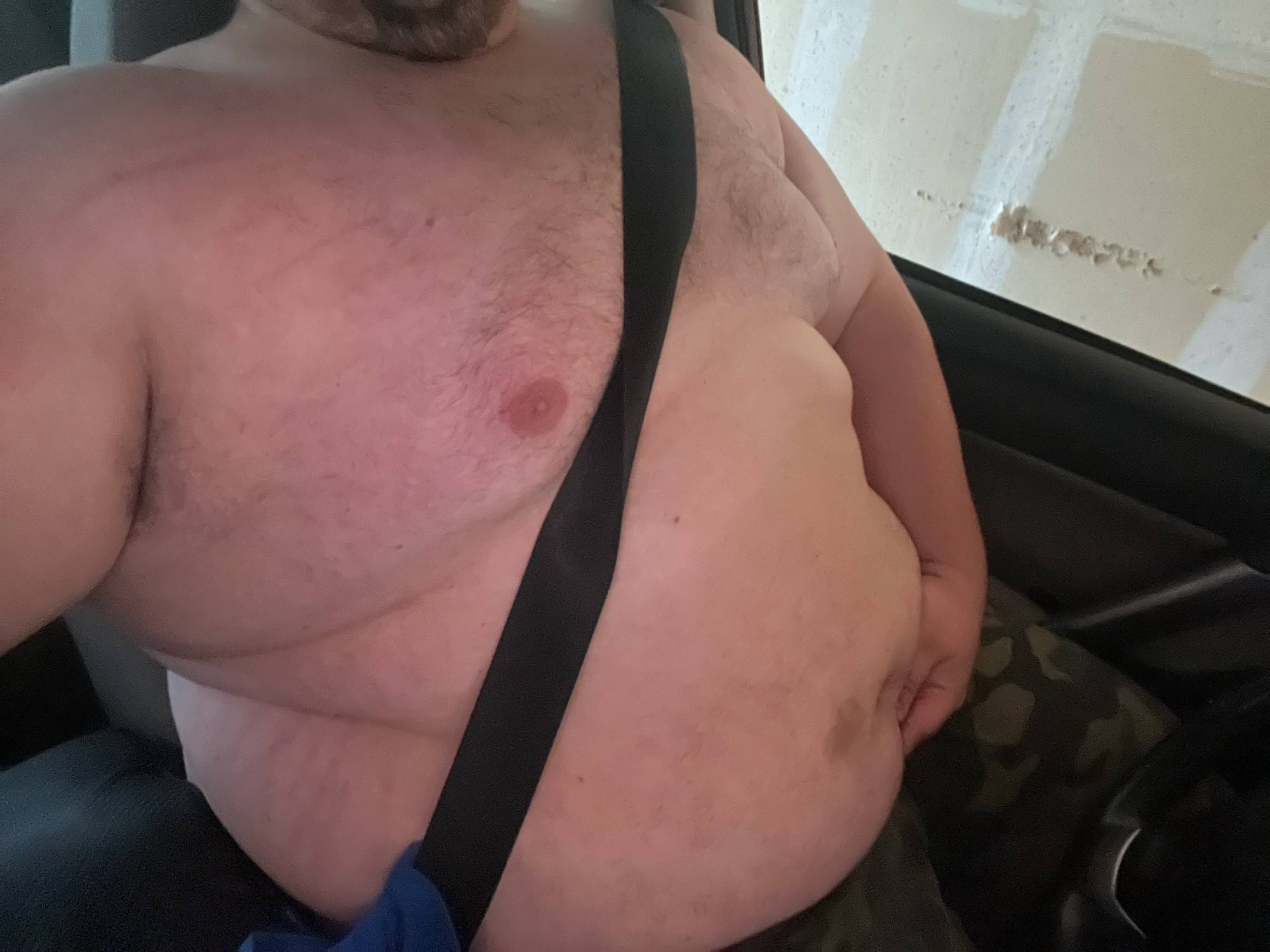 A large wet man shirtless in the car with a seatbealt on