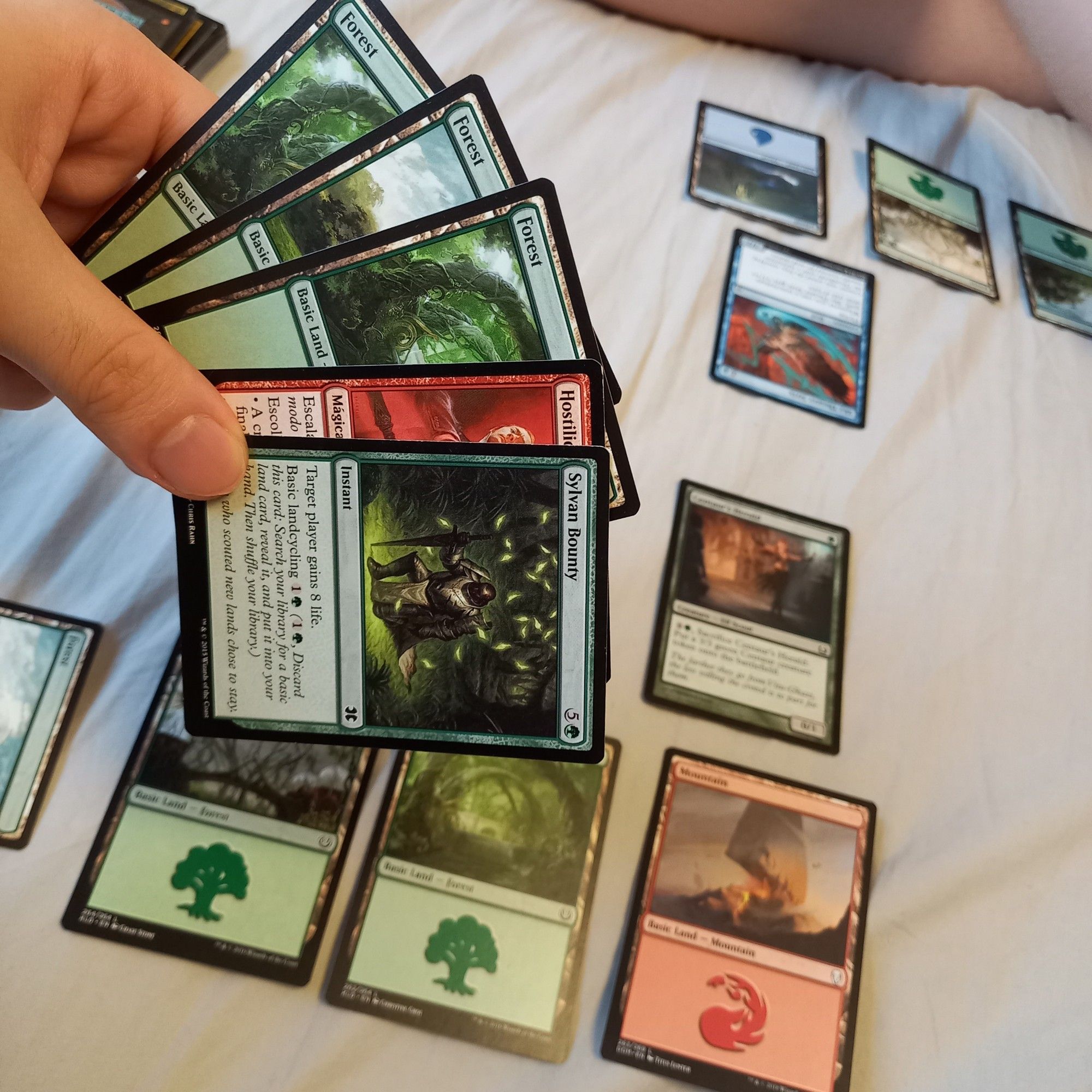 Mão segurando 4 cartas de Magic verdes e vermelhas. Ao fundo um campo de partida com terrenos verdes, vermelhos e azuis com criaturas verdes e azuis em campo.