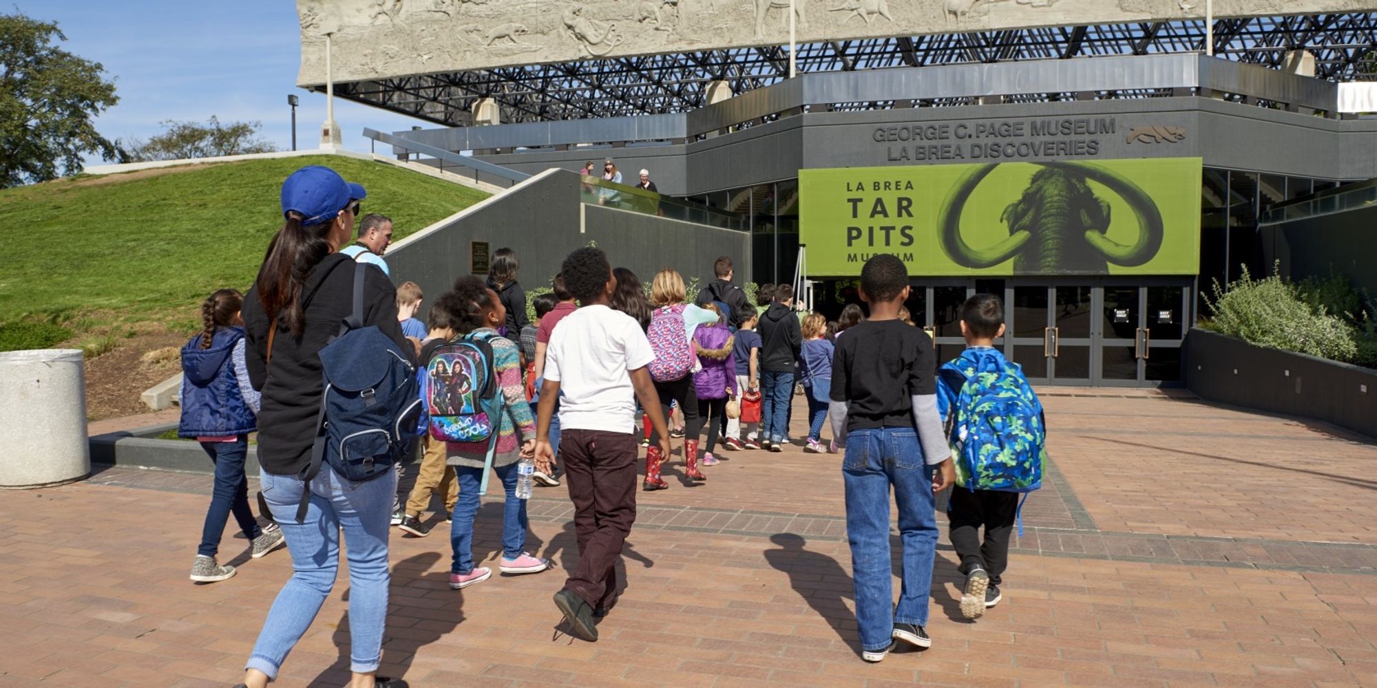 A group of children led by educators walk toward the entrance of La Brea Tar Pits & Museum.