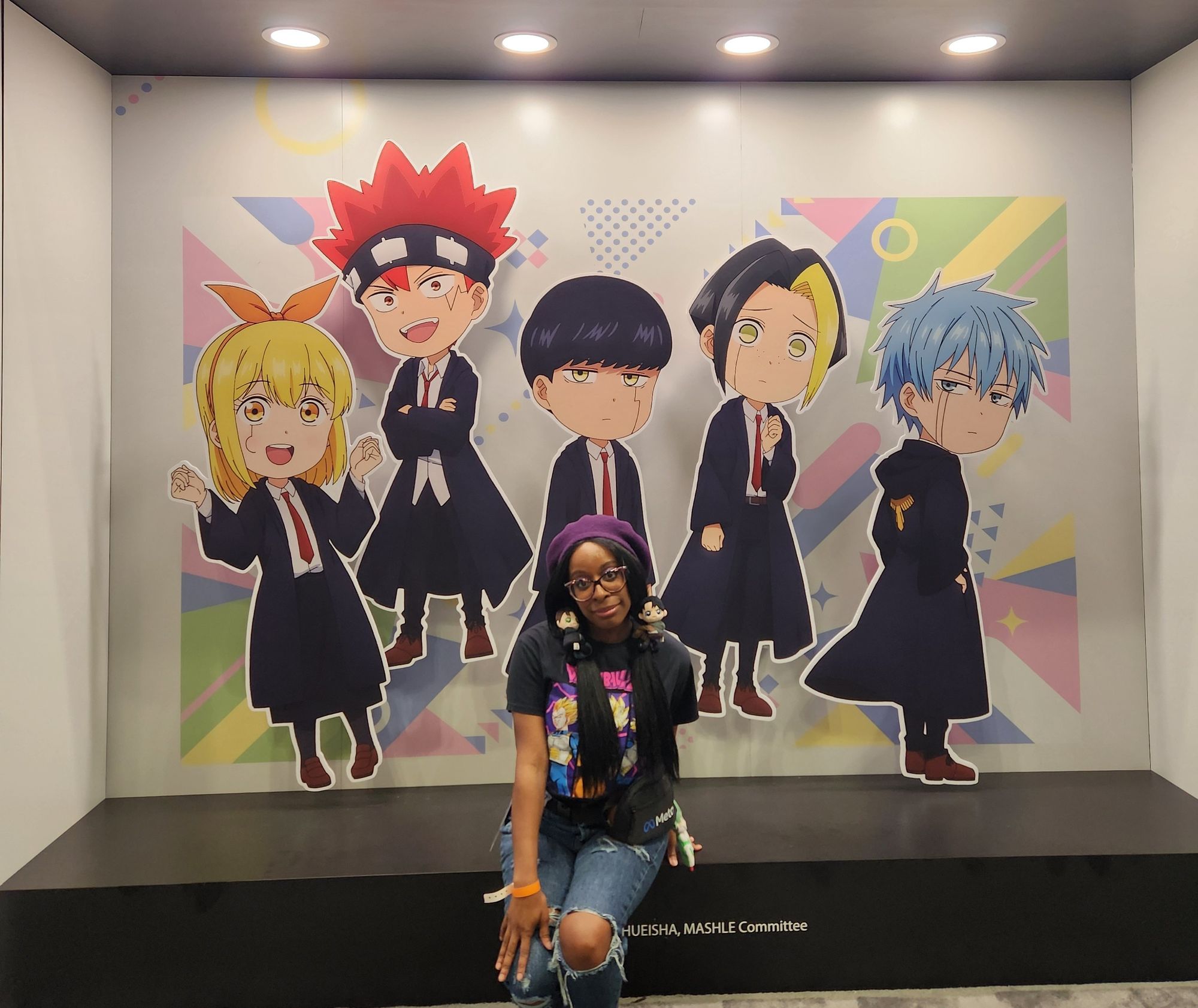 Woman sitting in front of a promotional image for the anime Mashle series. Woman is wearing a hat, glasses, and her hair is in pigtails. The image for Mashle are the 5 main characters in chibi mode