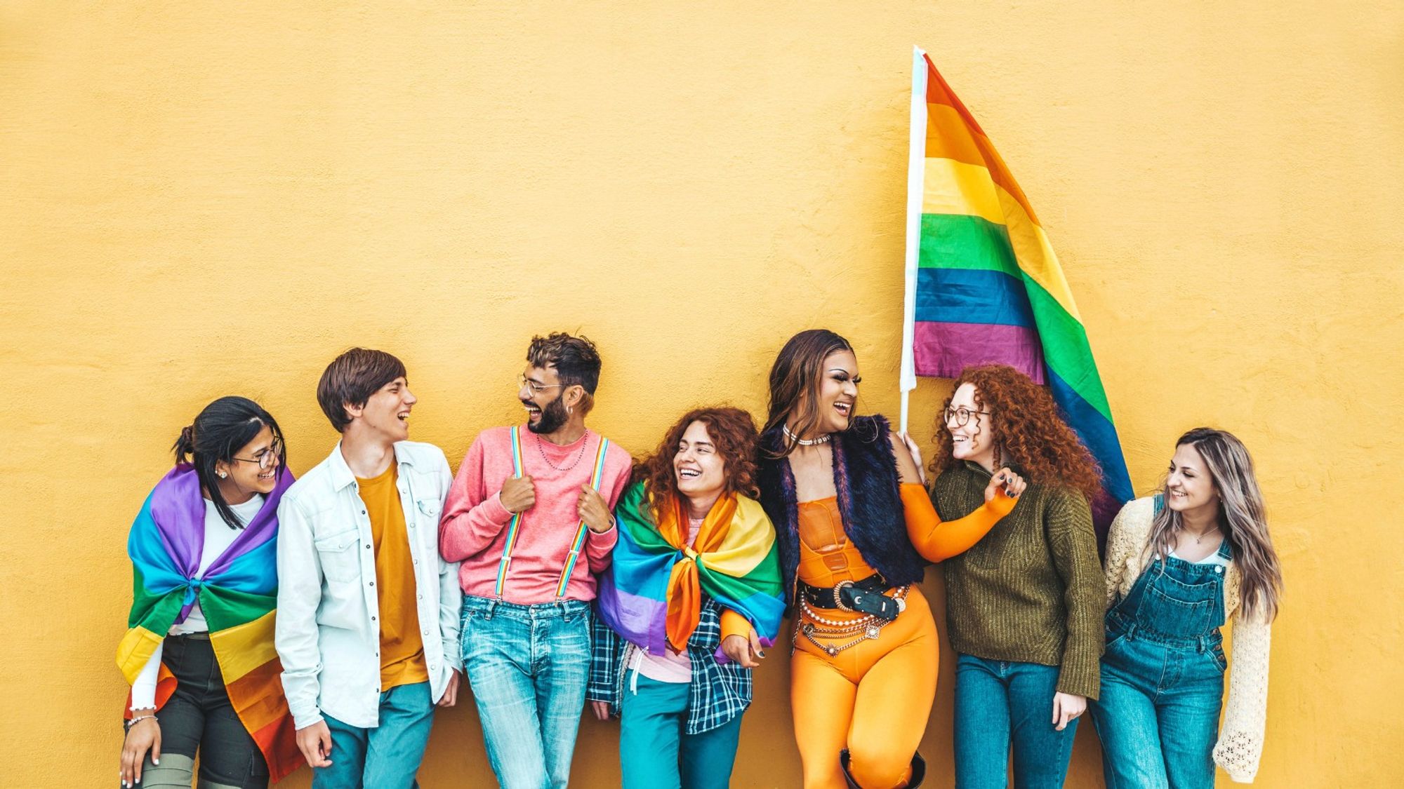 A photo of seven people draped in pride flags looking at each other with happiness.