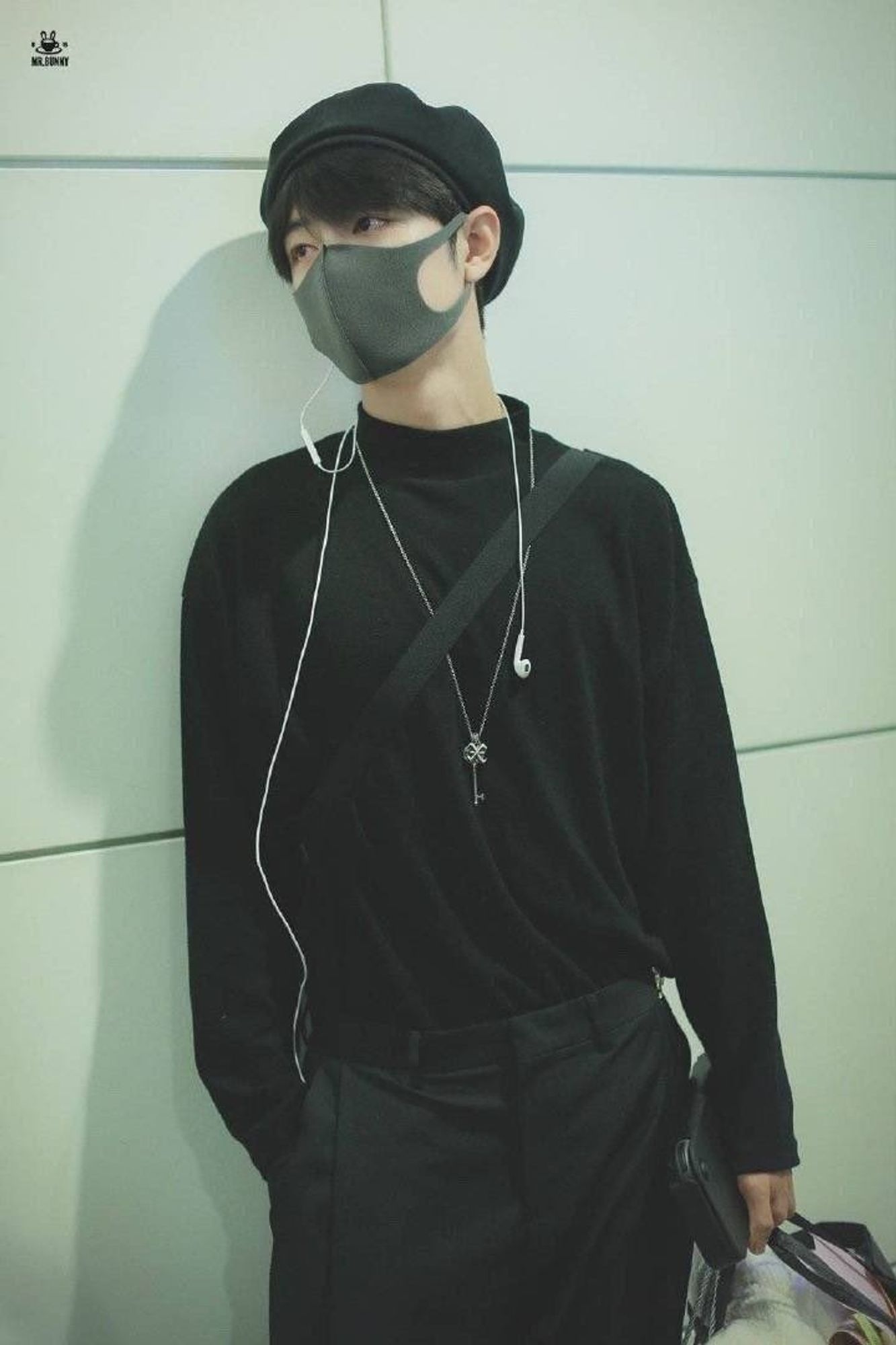 Xiao Zhan leaning against a wall at the airport. He's wearing all black, including a beret and a face mask, and holding a Nintendo Switch case.