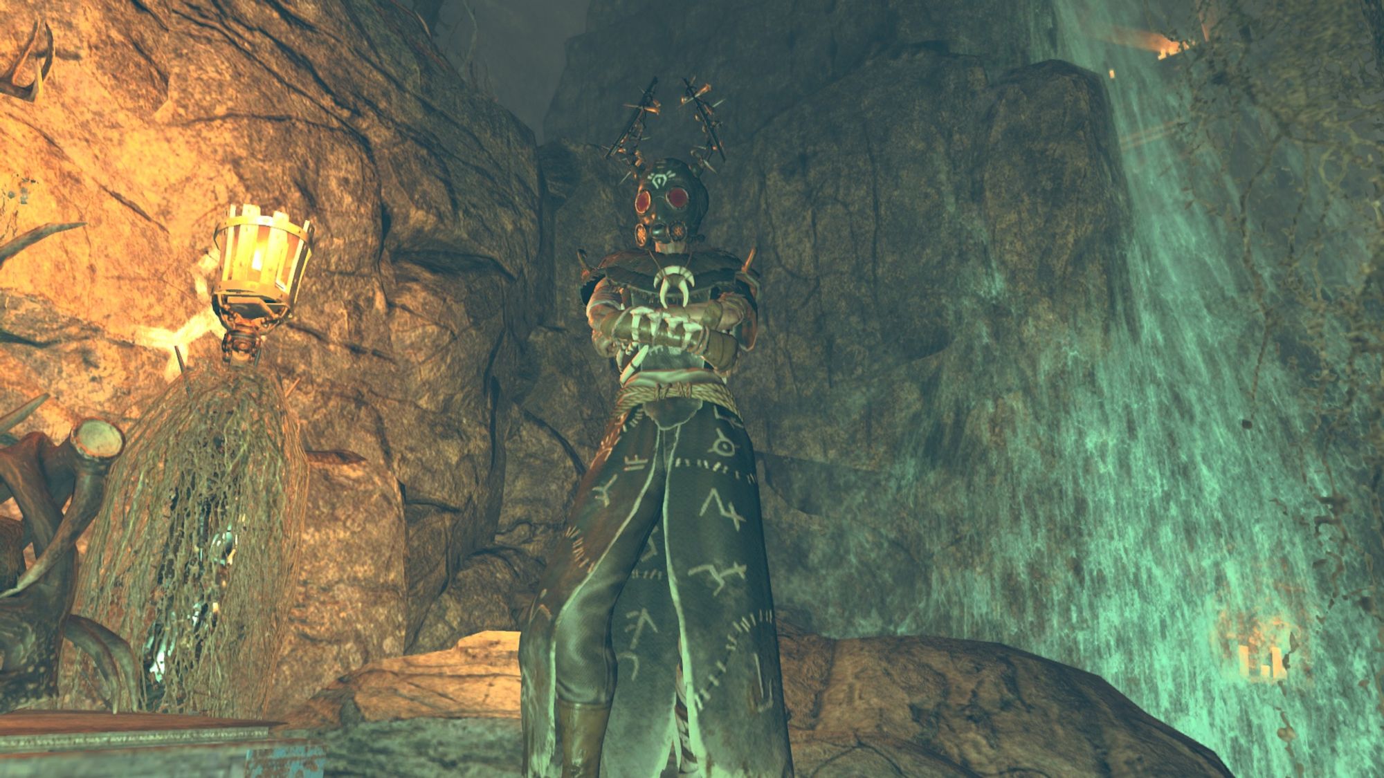 Screenshot from the game Fallout 76 focusing on the character Alex Guera, introduced in the Skyline Valley update. Alex is stood with arms crossed, looking of camera to the viewer’s left. Alex is wearing an ornate robed outfit with runic markings, a gas mask, and horned headwear.