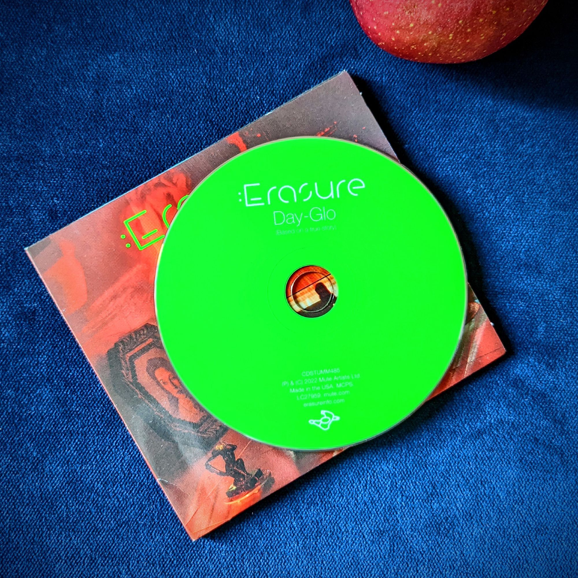 A bright neon green audio CD on a cardboard sleeve. Printed in white on the CD is “Day-Glo,” just below the artist’s name, which is Erasure. The CD and sleeve are on jewel-tone blue velvet with part of a red apple visible just in the frame.