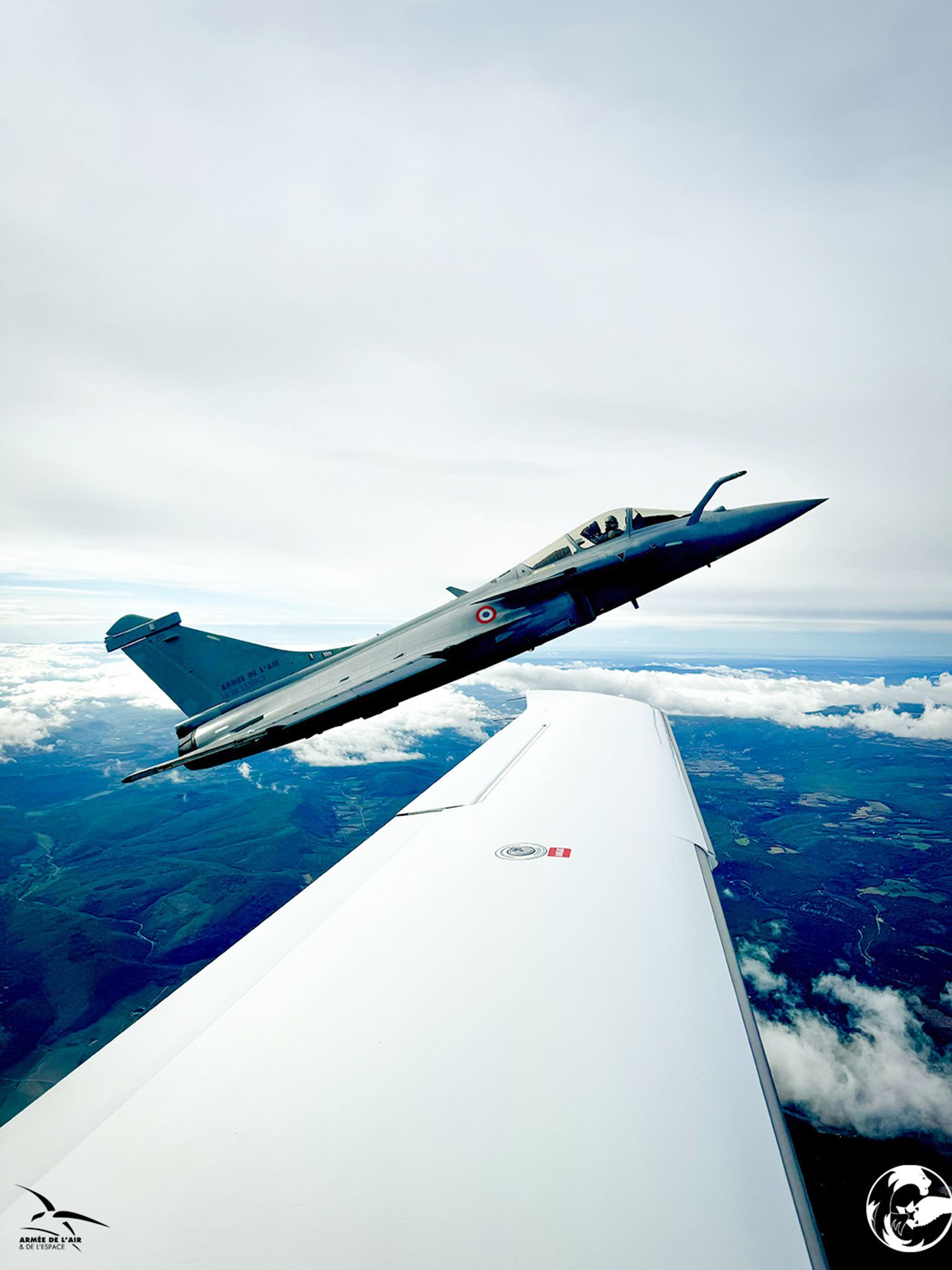 Au premier plan, l'aile d'un avion SR-22. Au second plan, un chasseur Rafale avec une forte incidence vole en formation rapprochée. A l'arrière-plan, un paysage de campagne partiellement recouverte de nuages bas. La couverture nuageuse en altitude est de 100 %.