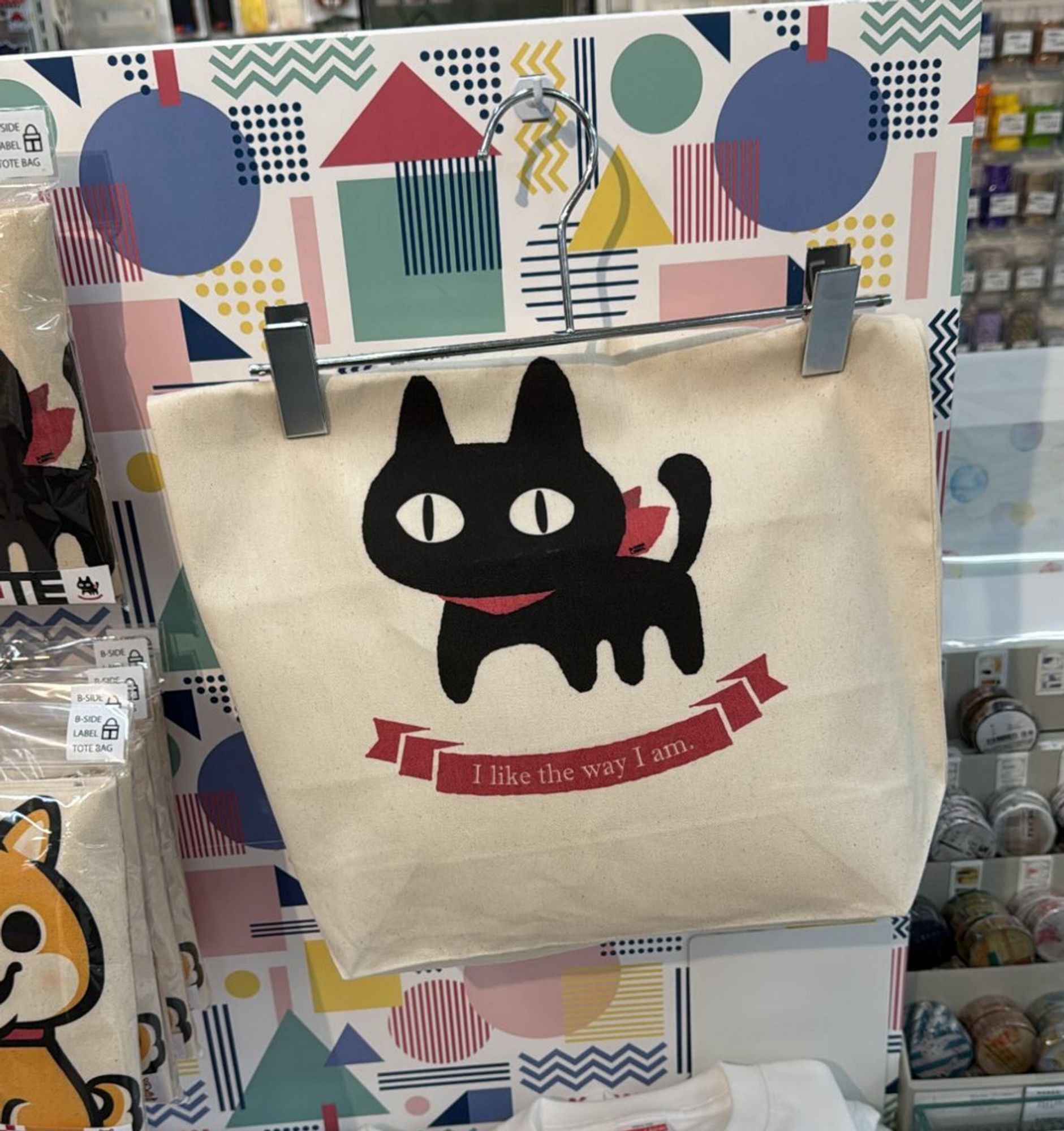 A tote bag with a black cat, wearing a red scarf with the message "I like the way I am" written below him.