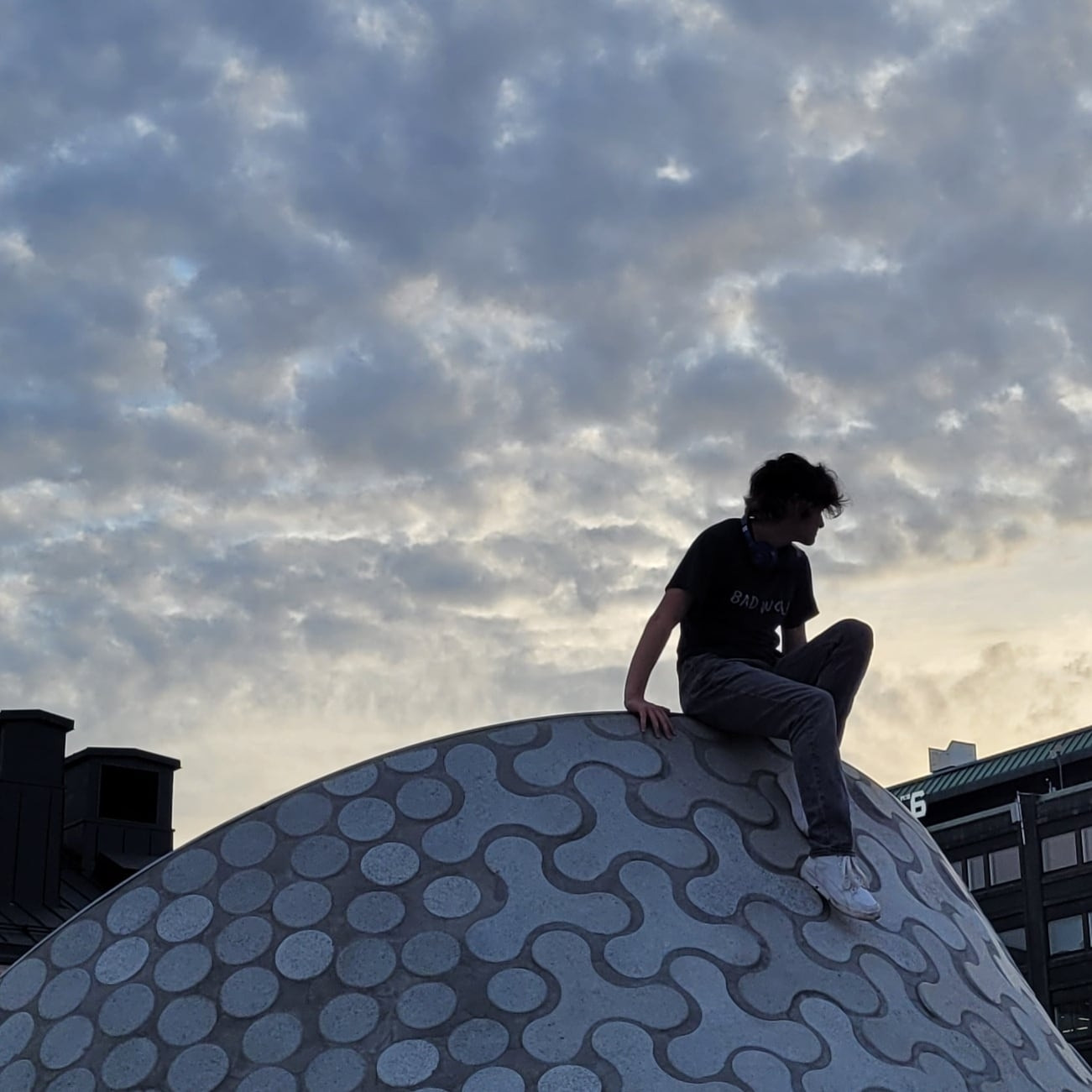 My 18 year old son in silhouette, sitting perched on top of a hill-like skylight in downtown Helsinki, with a clouded sunset sky behind him.