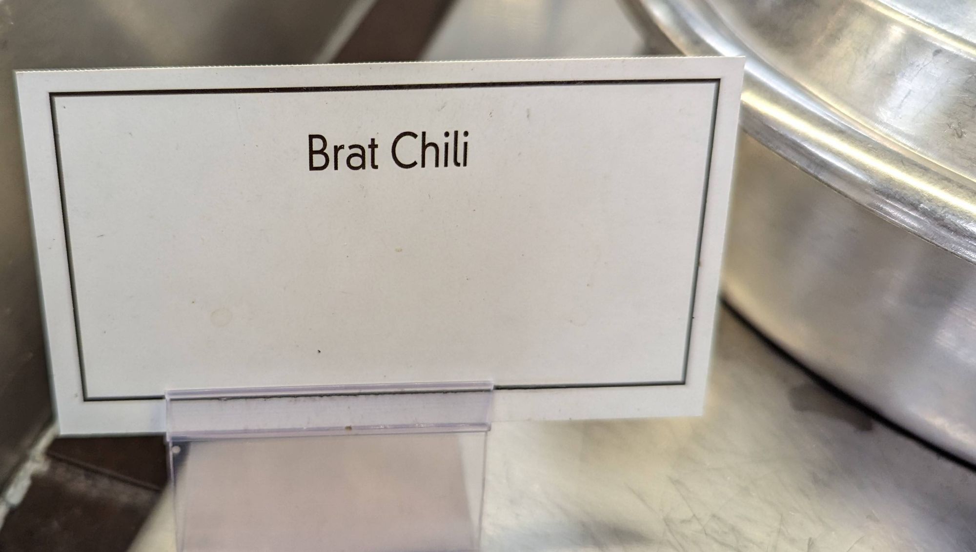 A paper card with the words "Brat Chili" held up by a plastic stand atop a metal surface. On the right in the background is part of a soup kettle lid.